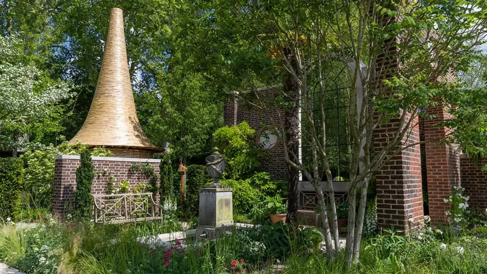 A contemporary show garden features a golden conical spire atop a brick plinth, a white decorative metal bench, and lush naturalistic planting with pink and white flowers.