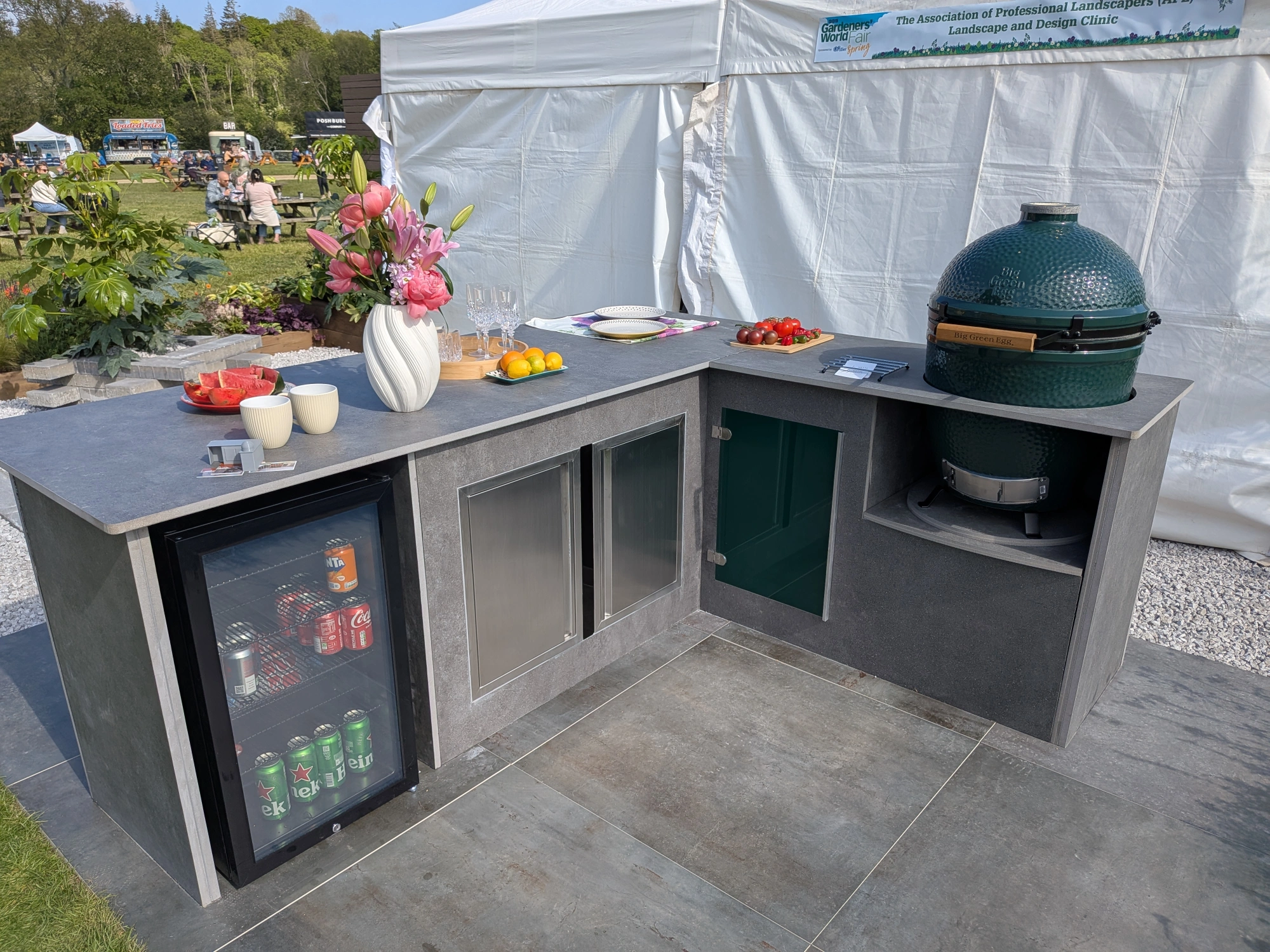 An L-shaped outdoor kitchen with dark grey porcelain countertops features an integrated fridge and a built-in Big Green Egg barbecue, styled for outdoor dining.