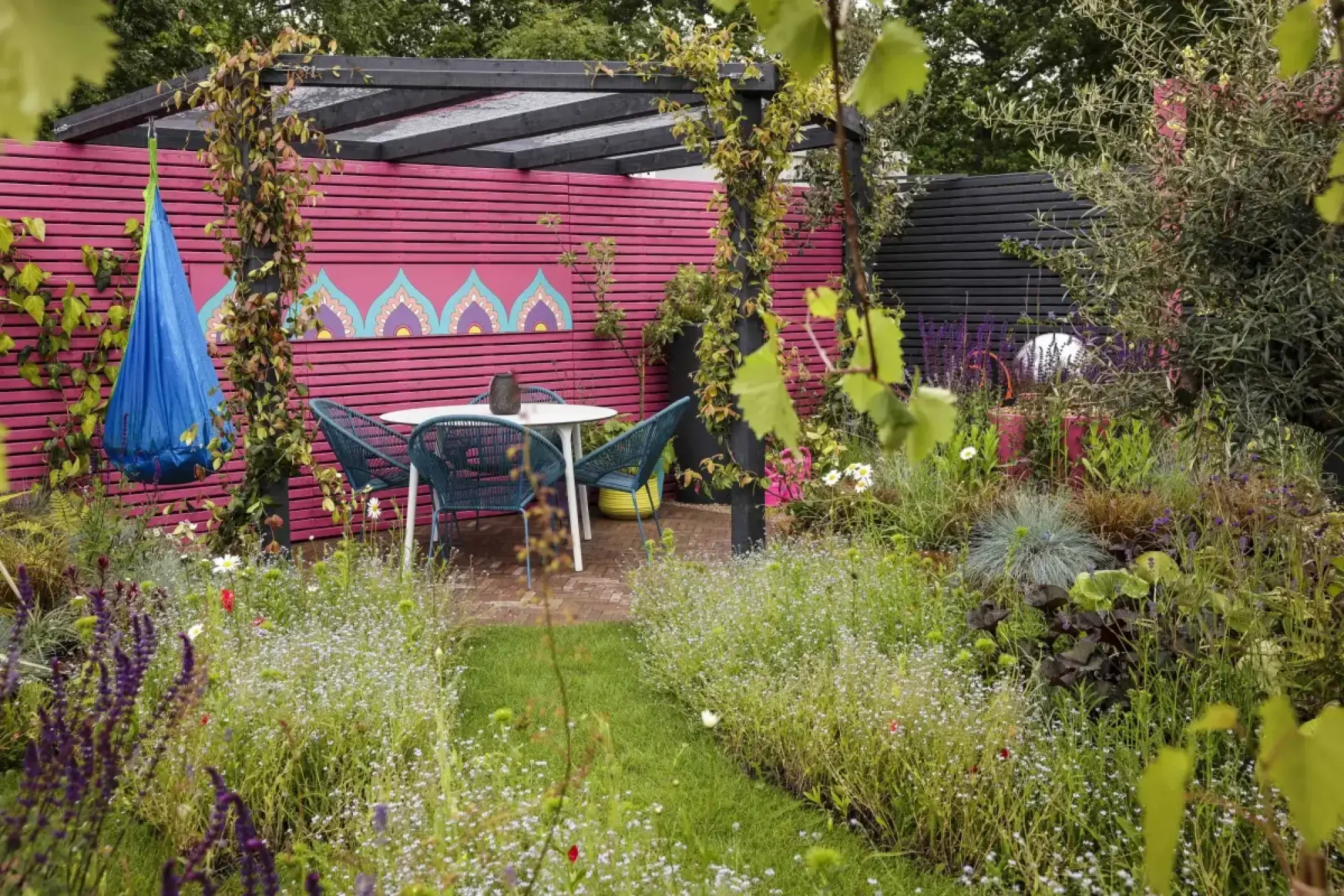 A vibrant wildlife garden features a hot pink fence with a mandala, a black pergola, a blue hanging swing, and a lawn path to a patio with teal chairs.