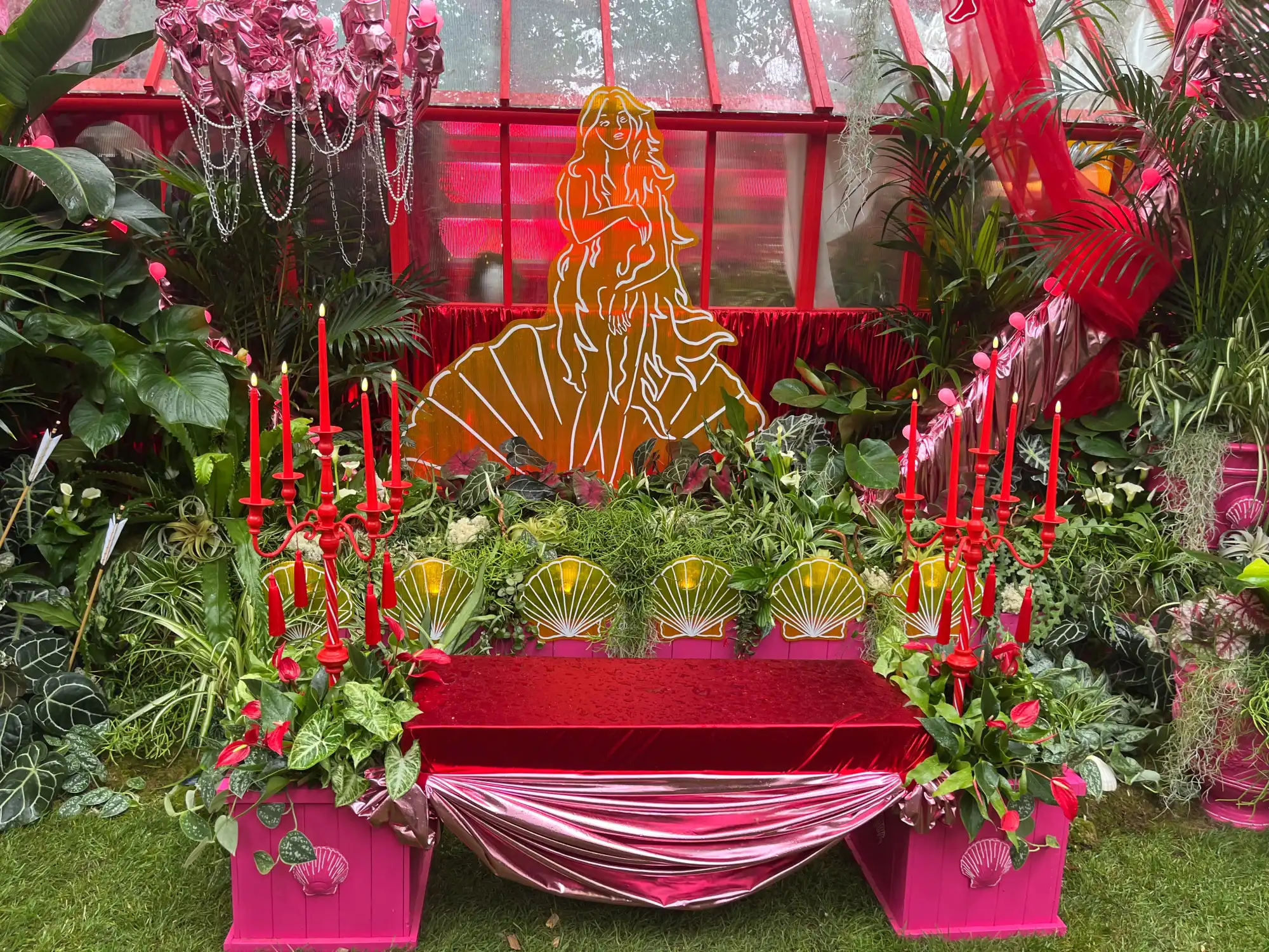 An opulent houseplant studio featuring a lit Aphrodite sculpture, a red velvet bench, and lush tropical plants surrounded by red and pink decor.