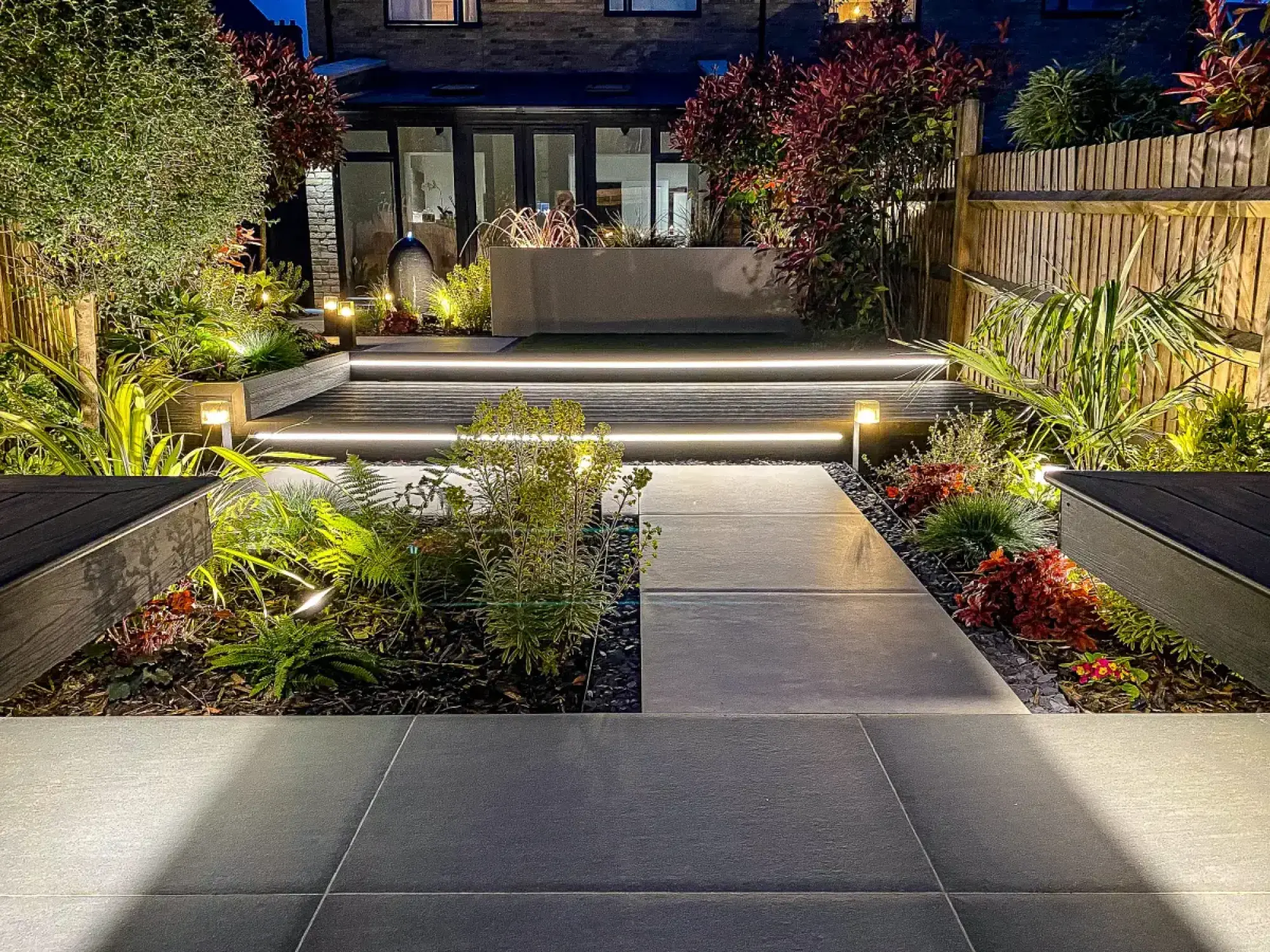 A contemporary garden at night with illuminated timber steps, dark grey paving, a ceramic water feature, and lush mixed planting.