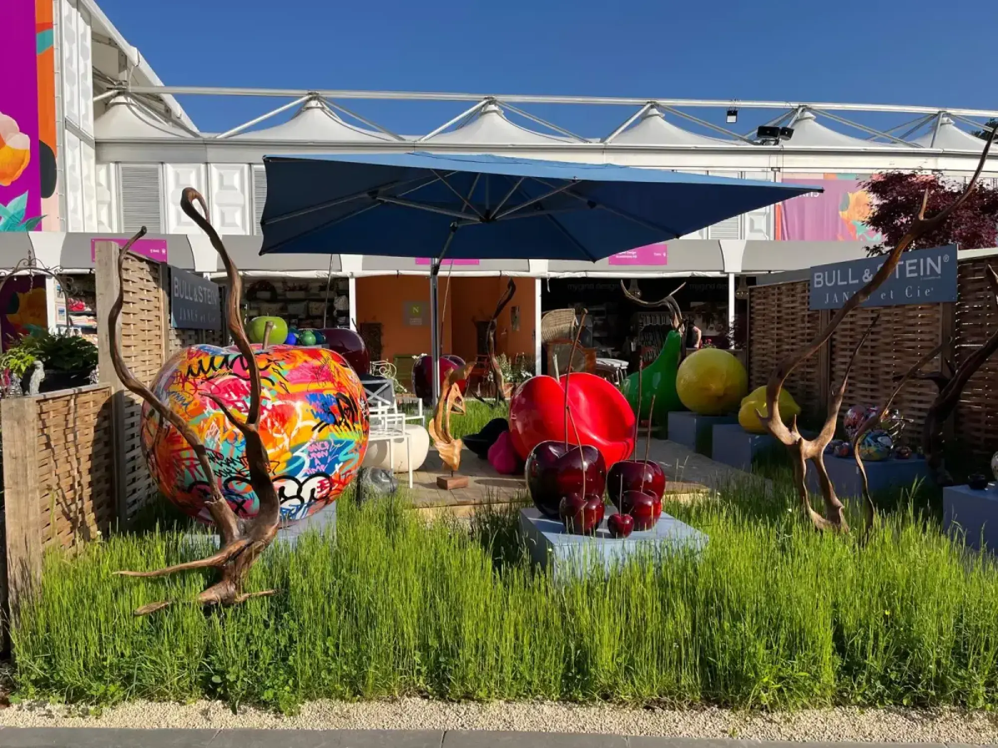 A vibrant trade stand features giant, colourful fruit sculptures, including red cherries and a graffiti-painted apple, displayed on grey plinths amidst long green grass and shaded by a large blue parasol.