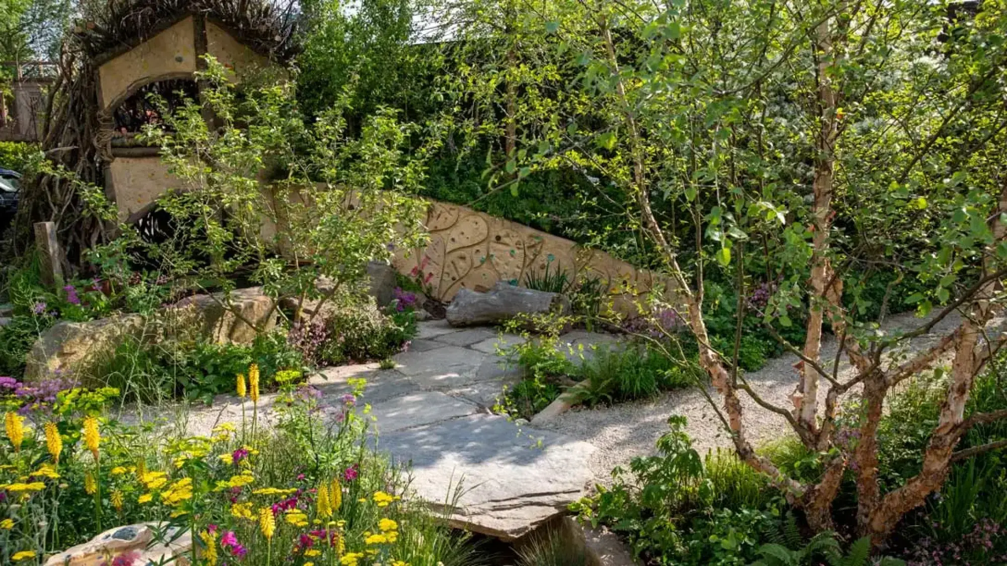 A naturalistic children's garden features a monster's mouth den, a sculpted wall, flagstone paths, and abundant planting with yellow and purple flowers.