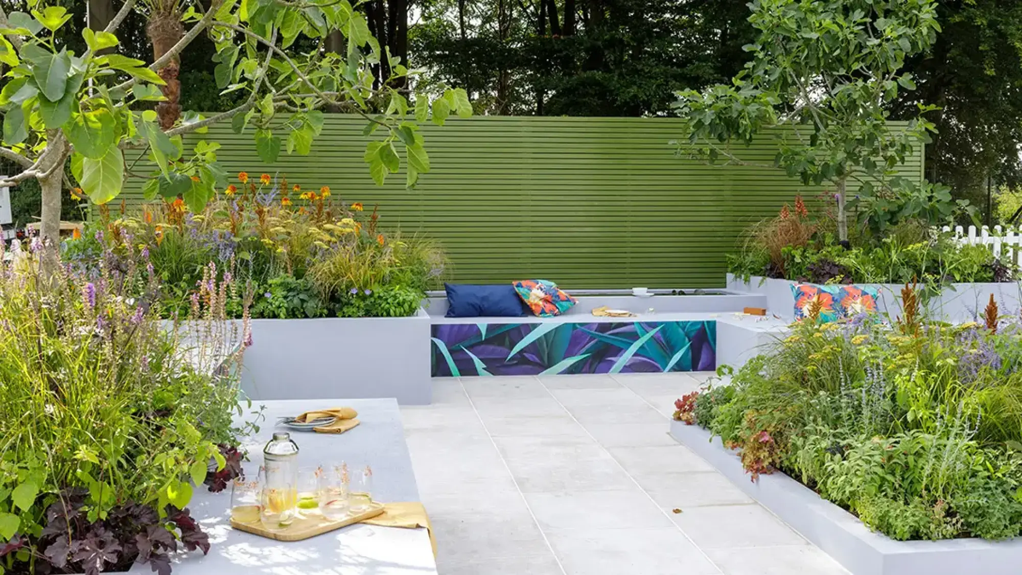 A modern garden with grey paving, colourful planters, an integrated bench with a purple and teal botanical art panel, and a striking green slatted fence.