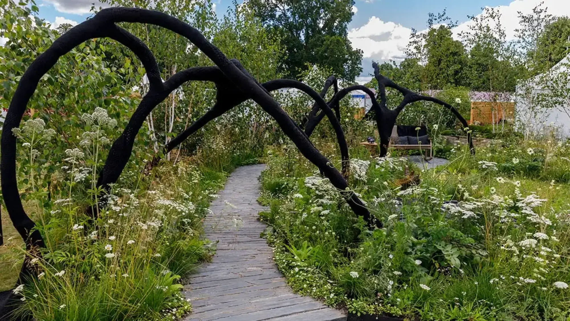 A naturalistic show garden with dramatic black charred arching sculptures, a winding grey path, and abundant white wildflowers among green grasses.
