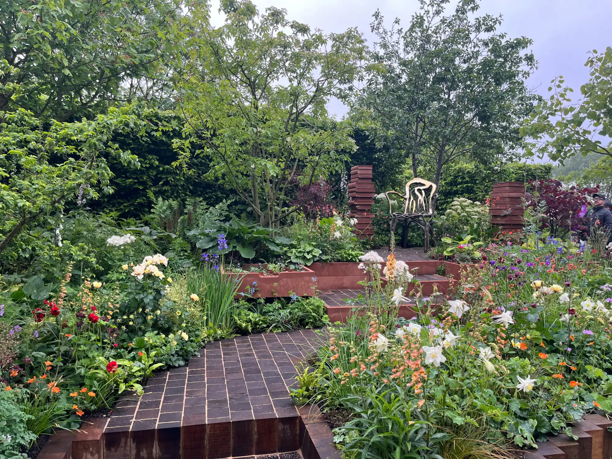 A lush, terraced garden featuring a gold sculptural chair, stacked terracotta pots, and abundant mixed perennial flowers in a spectrum of colours.