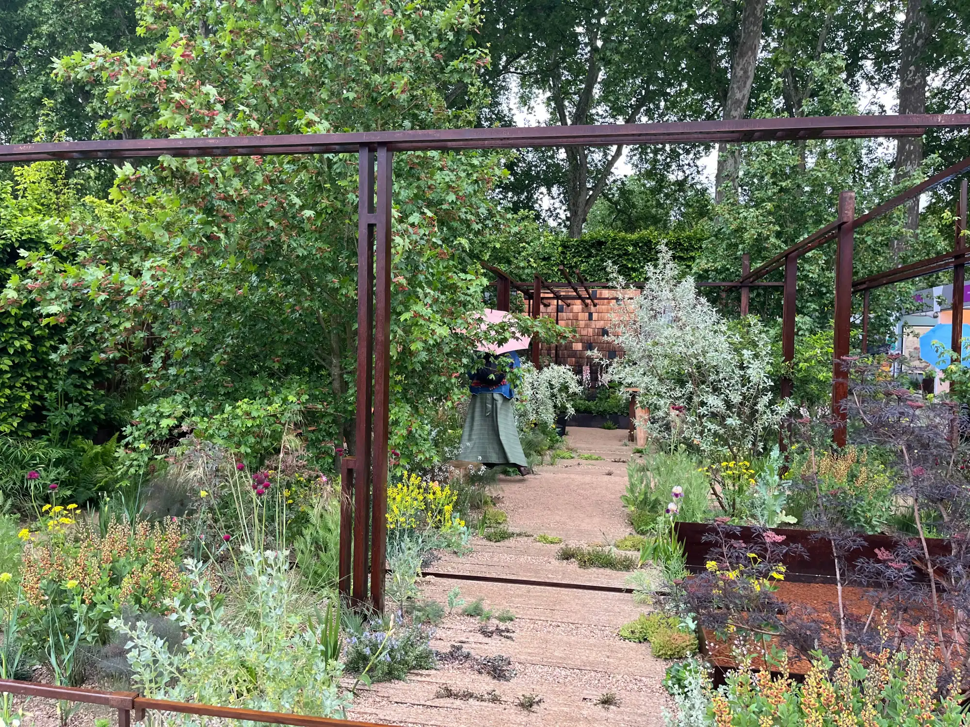 A naturalistic show garden features a timber path leading through lush mixed planting and a rust-coloured metal pergola to a shingle-clad structure.