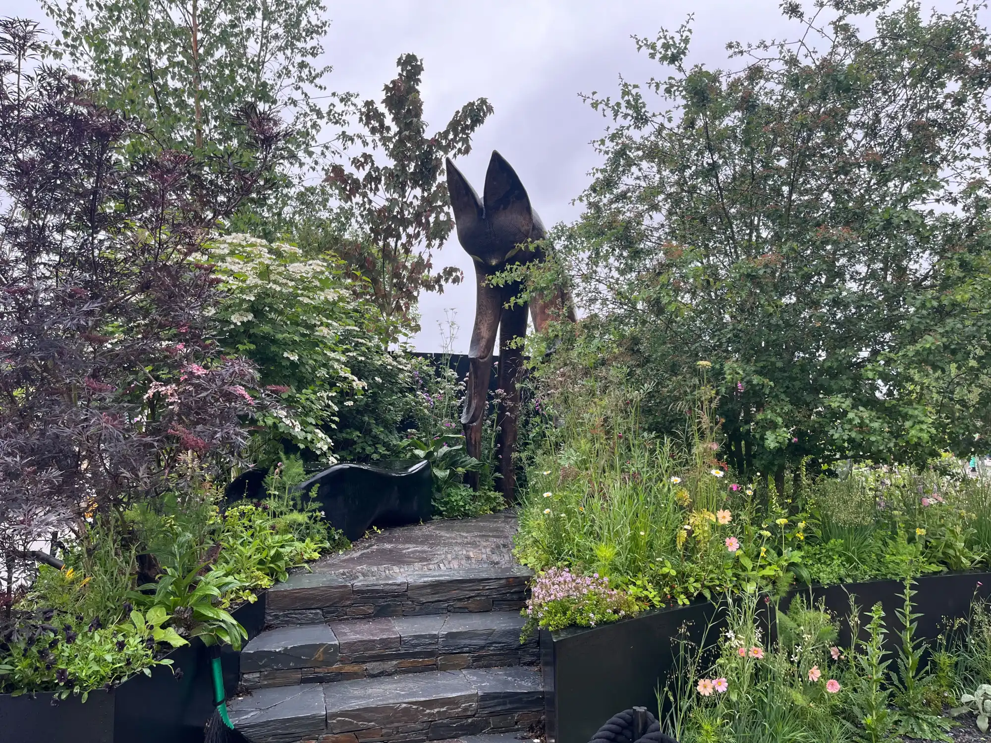 A naturalistic garden features layered slate steps leading to a tall, dark abstract bat ear sculpture, surrounded by lush planting with dark purple foliage, white flowers, and delicate pink and white wildflowers in black planters.