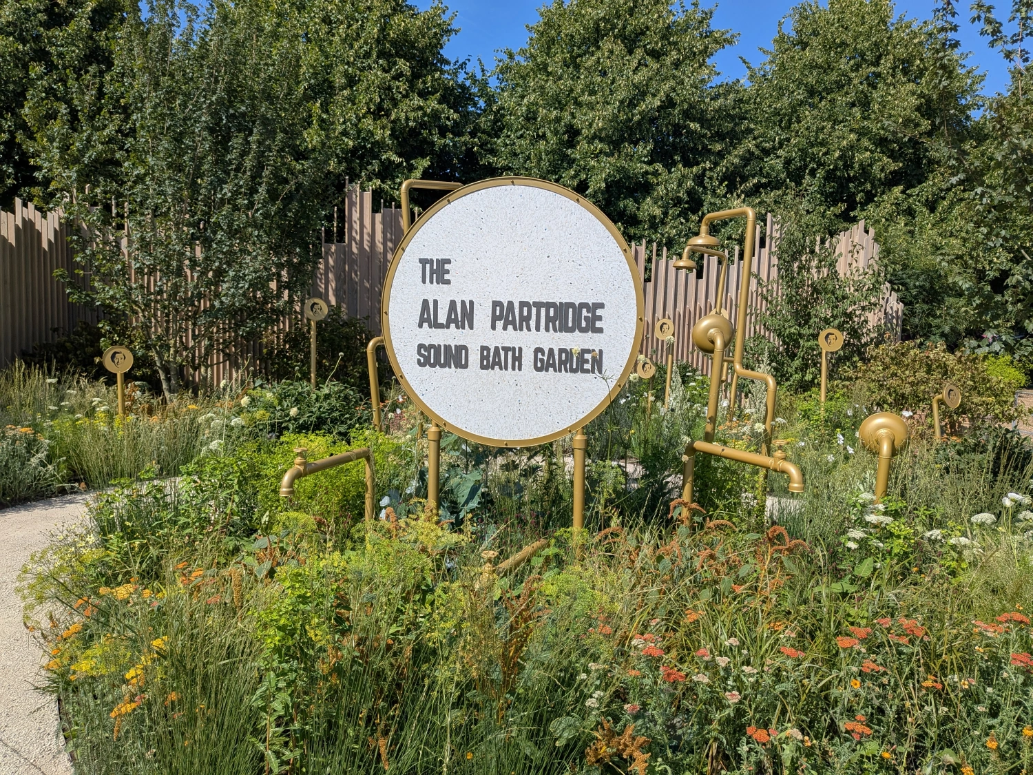 A whimsical show garden features a white circular sign and gold pipe structures like taps and shower heads, surrounded by naturalistic green and colourful planting.