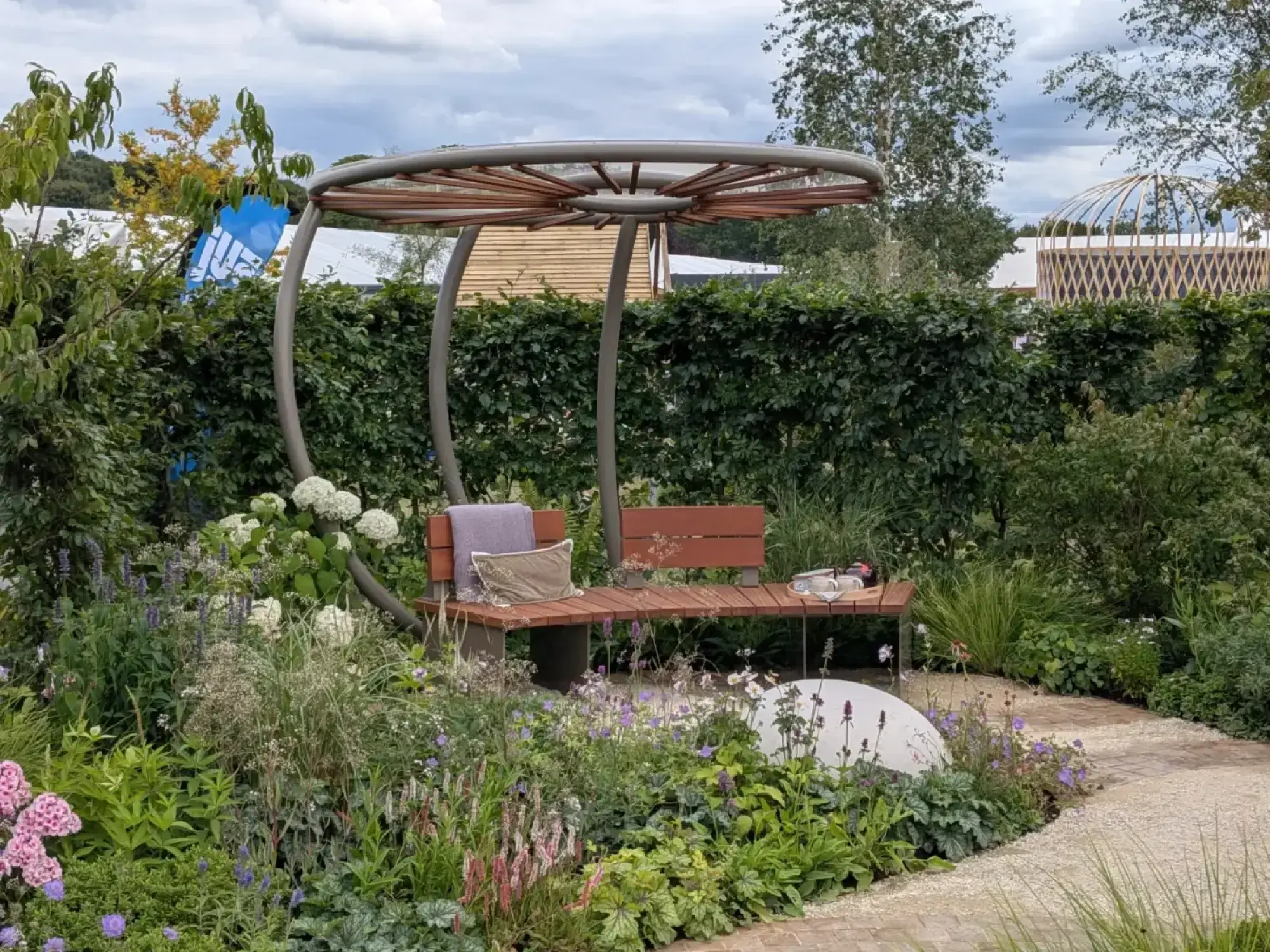 A tranquil show garden features a curved metal pergola over a wooden slatted bench, surrounded by abundant naturalistic planting including white hydrangeas, pink phlox, and purple flowers, with a gravel path.