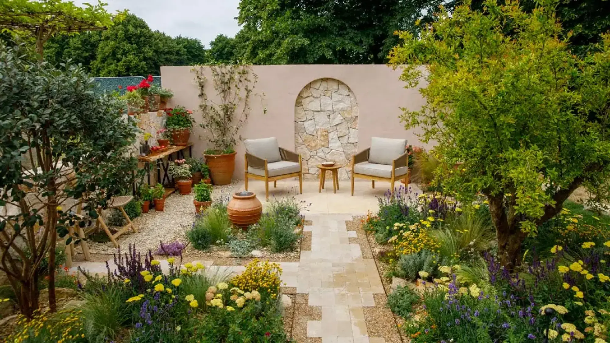 A Mediterranean garden features a pink wall with an arched stone niche, armchairs, a path, and lush beds of purple and yellow flowers.
