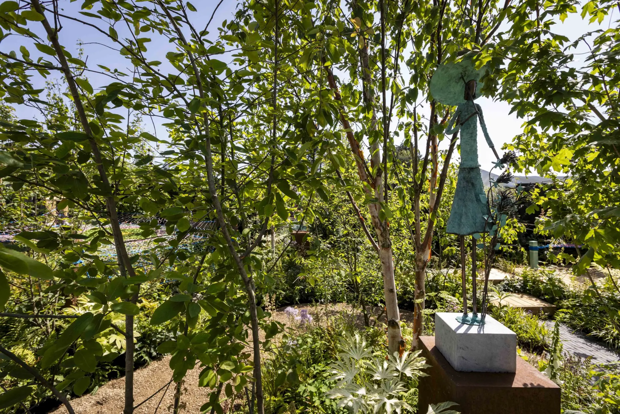 A naturalistic garden features a teal patina sculpture of a woman with a wide-brimmed hat holding a bouquet, amidst silver birch trees, lush green foliage, purple flowers, and a winding gravel path.
