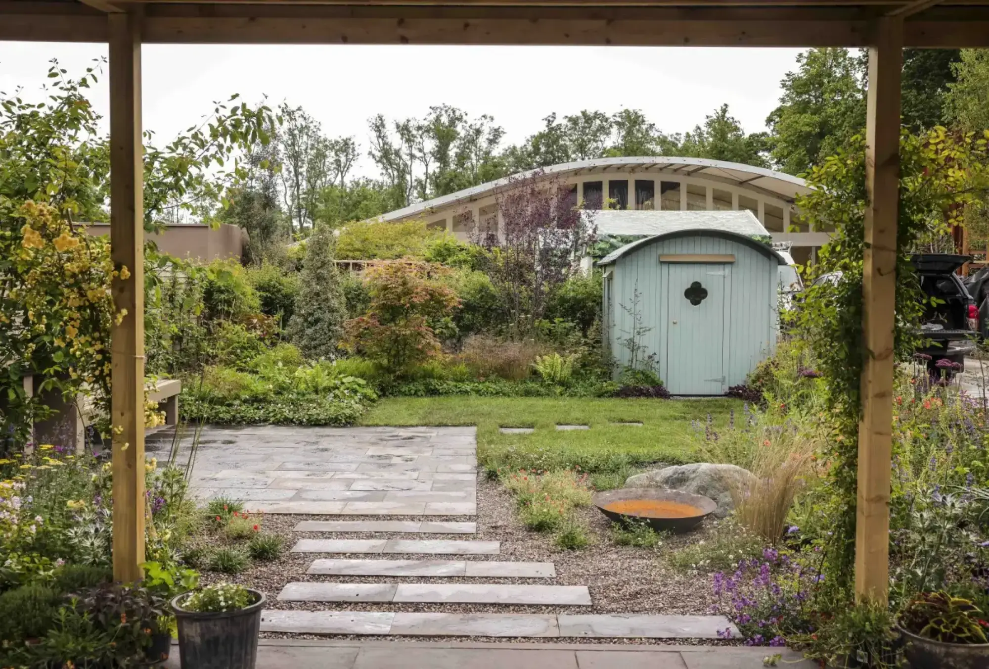 A lush, informal feature garden with a light blue quonset hut style shed, a rusted fire pit, and a grey flagstone patio with stepping stones leading through mixed herbaceous borders.