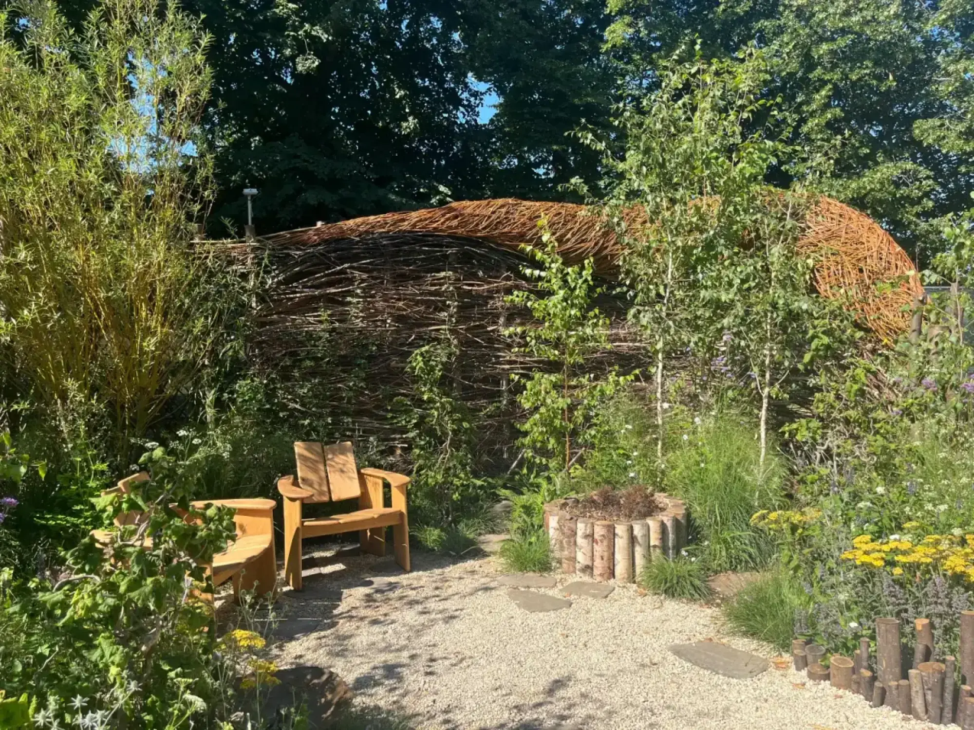 A naturalistic woodland garden with a large, organic woven branch structure, two wooden chairs, and wild planting with grasses, yellow and purple flowers.