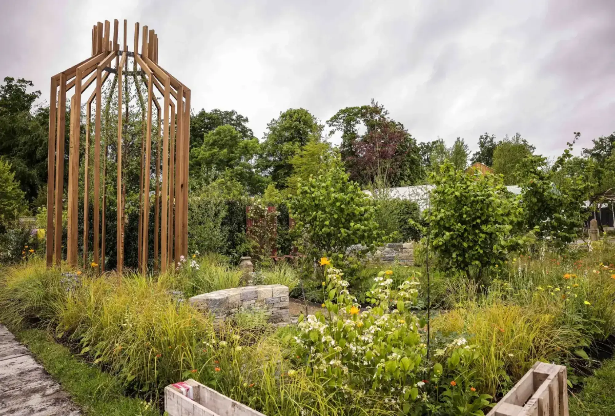 A large, naturalistic show garden features a tall, open timber structure enclosing a birch tree, surrounded by diverse ornamental grasses, flowering shrubs, and stacked stone walls.