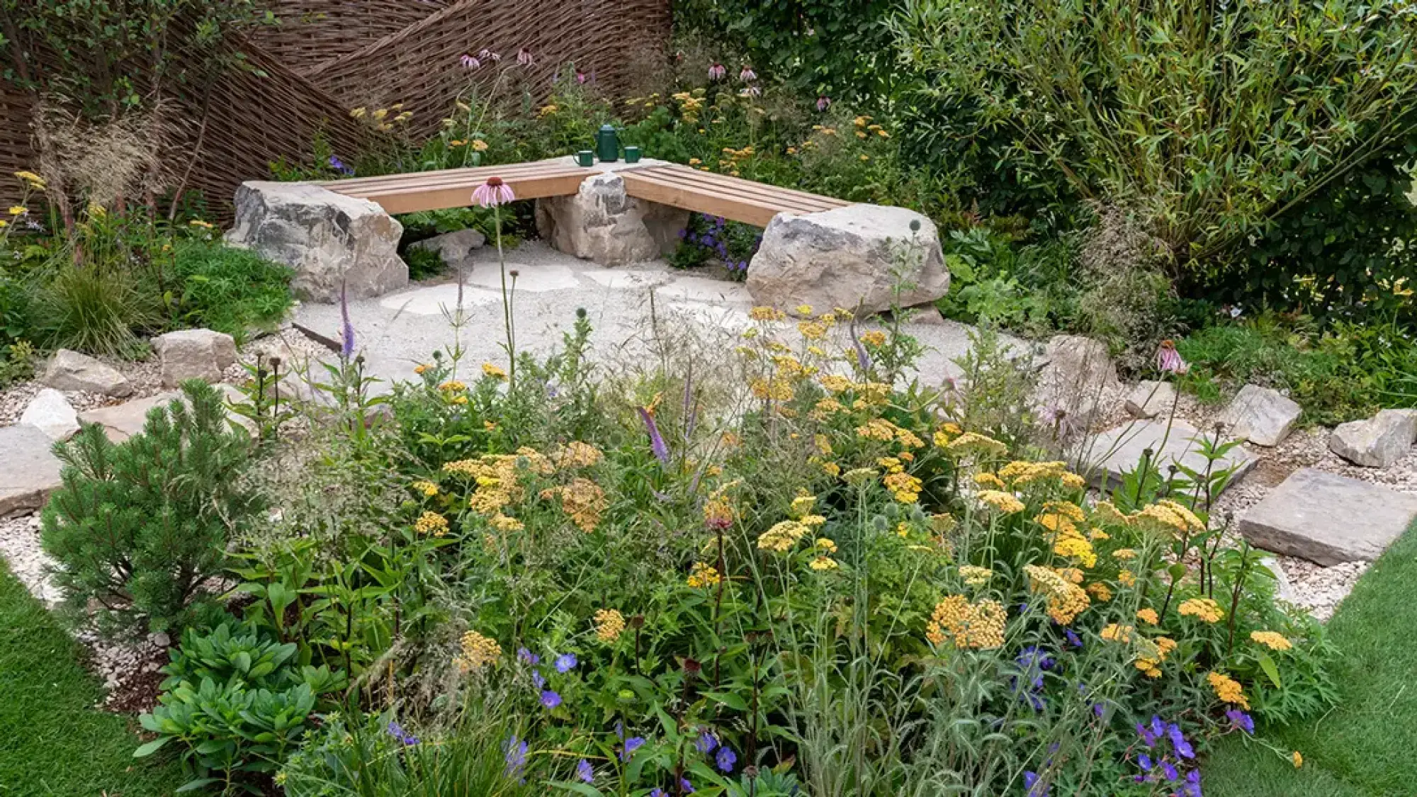 A naturalistic garden featuring a timber bench resting on large stone blocks, surrounded by abundant yellow, purple, and green wildflowers and grasses, with a gravel path and stone stepping stones.