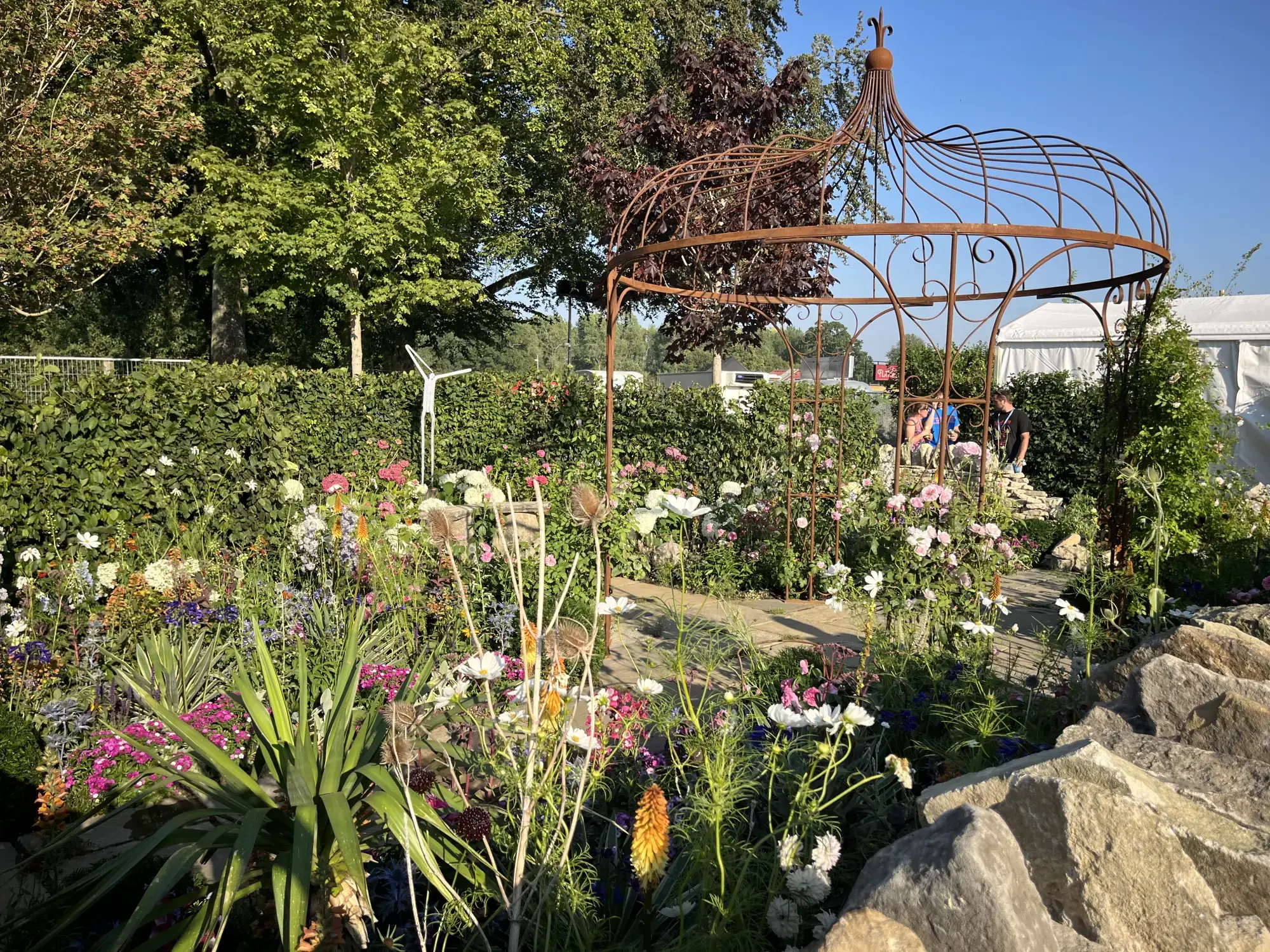 A rustic artisan garden features a large rusted metal gazebo surrounded by abundant mixed flowers, natural boulders, and a winding stone path.