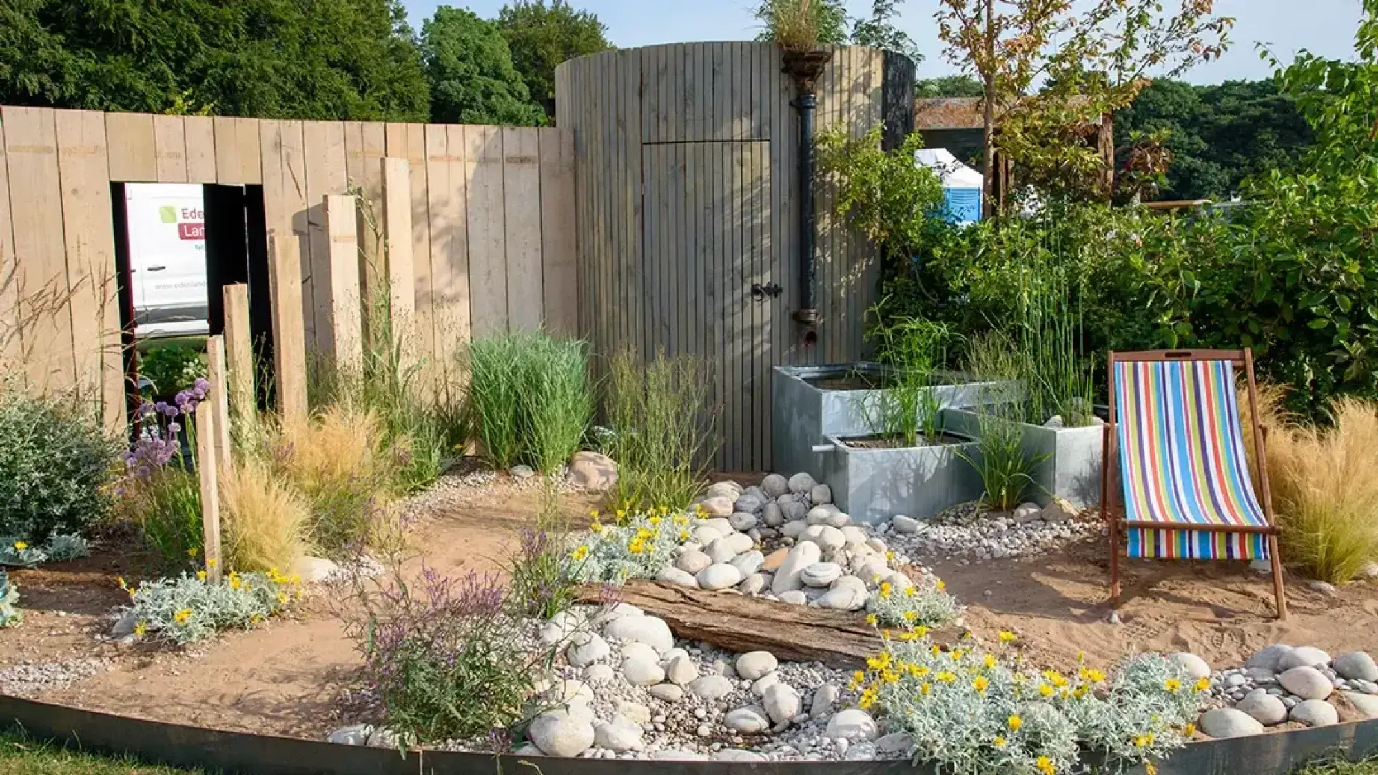 A relaxed coastal garden featuring a round timber building, tiered metal planters, a shingle beach, a striped deck chair, and naturalistic plants.
