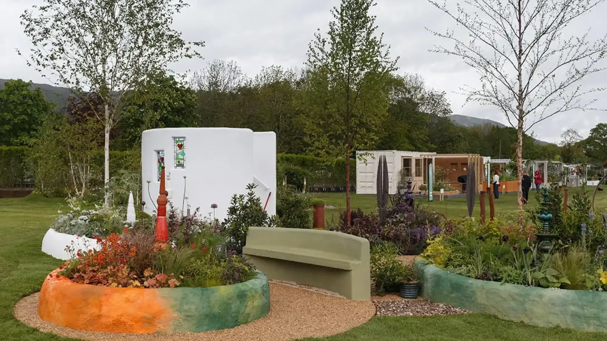 A contemporary show garden with a white curvilinear building, heart-motif stained glass, curved orange and green planters, a matching green bench, and mixed colourful perennials.