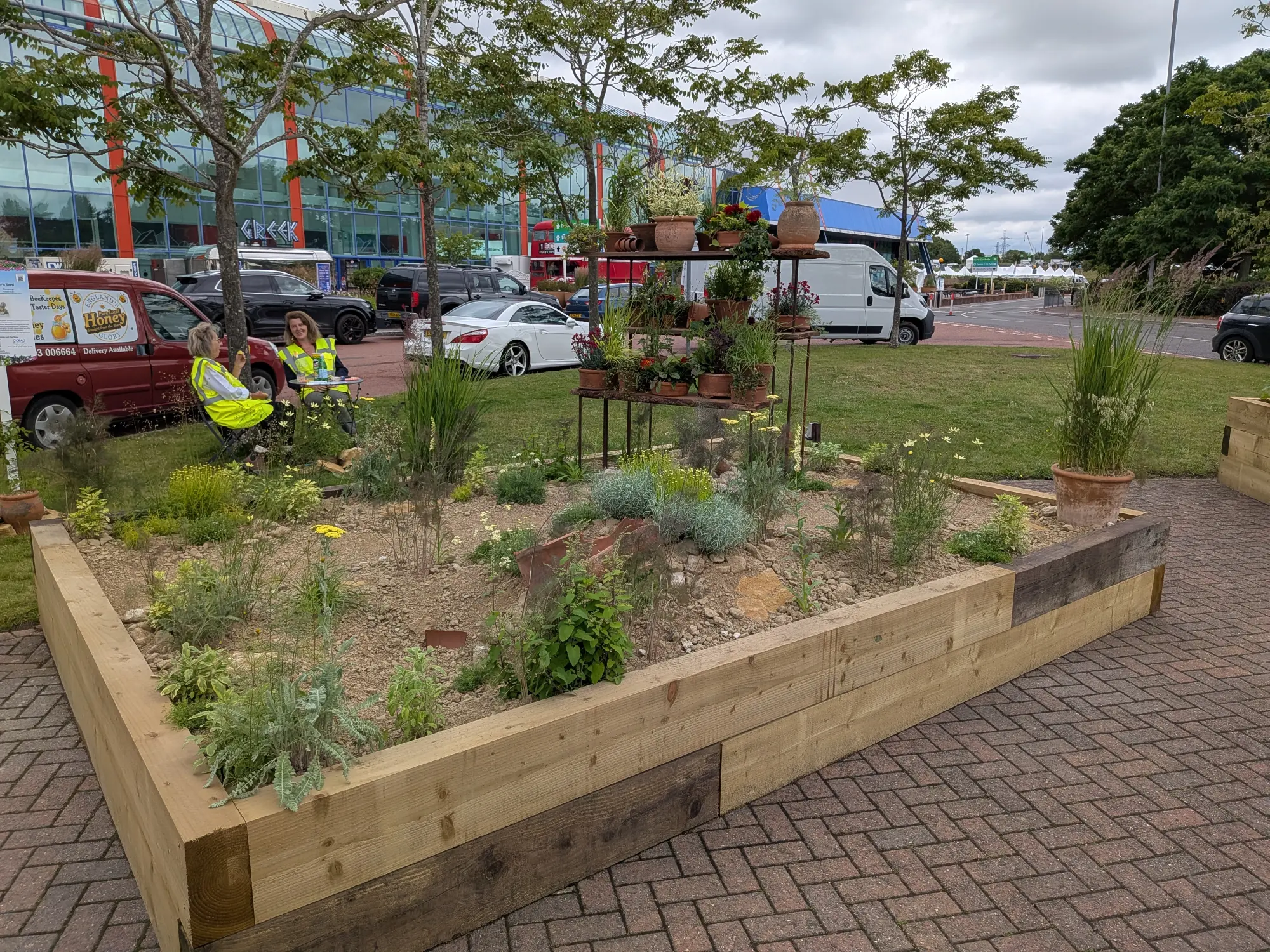 A rustic raised timber border holds a multi-tiered metal plant stand, terracotta pots, and drought-tolerant plants among scattered broken pottery.