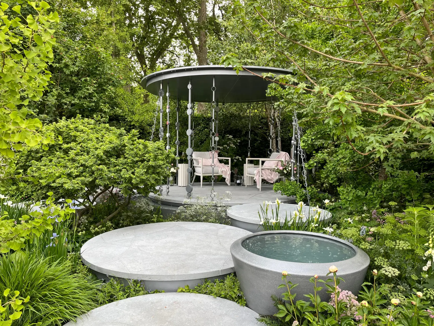 A contemporary garden featuring a circular grey pavilion with decorative chains, set among lush greenery, with round grey platforms and a stone water basin.