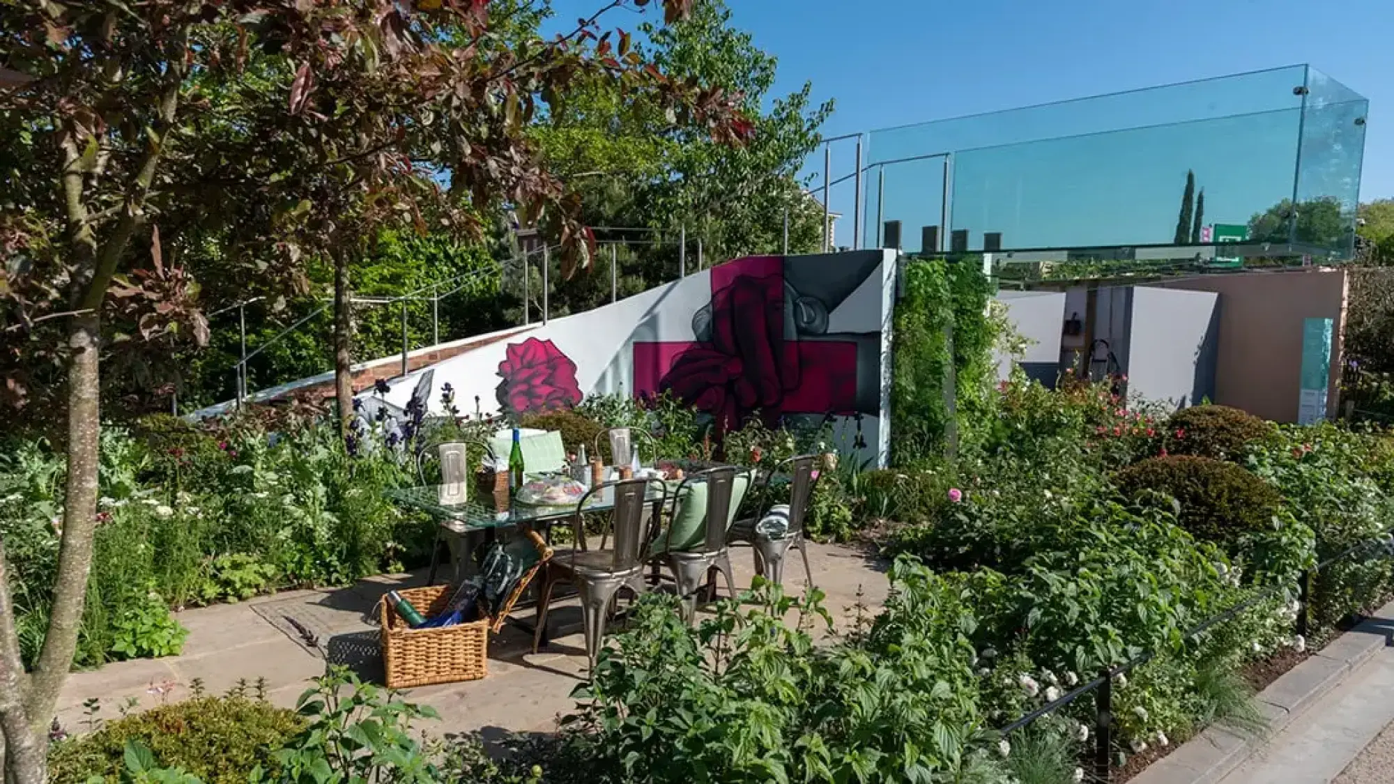 A contemporary sanctuary features a paved dining patio, lush plants, and a striking curved wall with a red and grey mural, topped by clear glass.