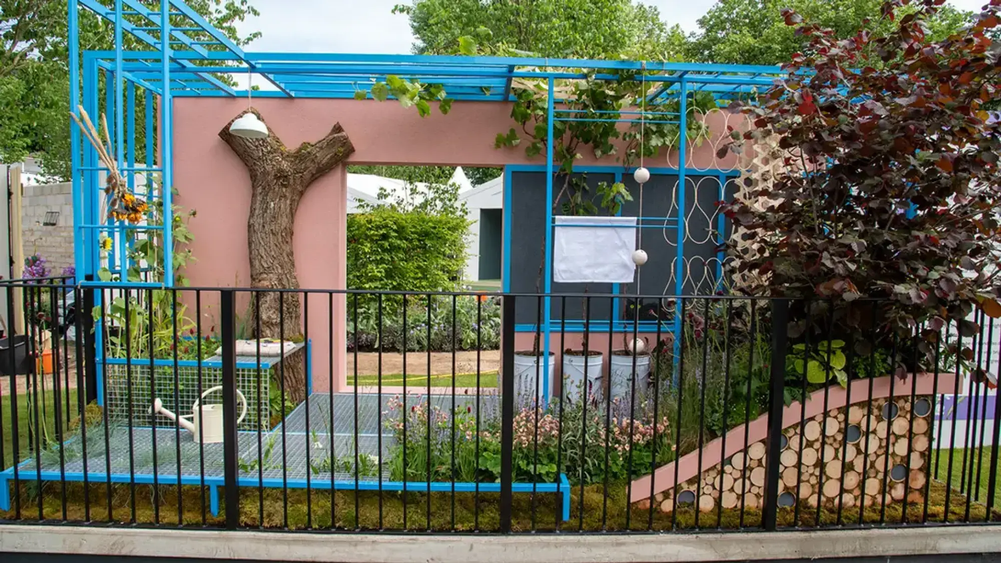 A modern balcony garden features a bright blue metal pergola, a pink wall with an integrated large tree trunk, and a curved wall made of wood slices.