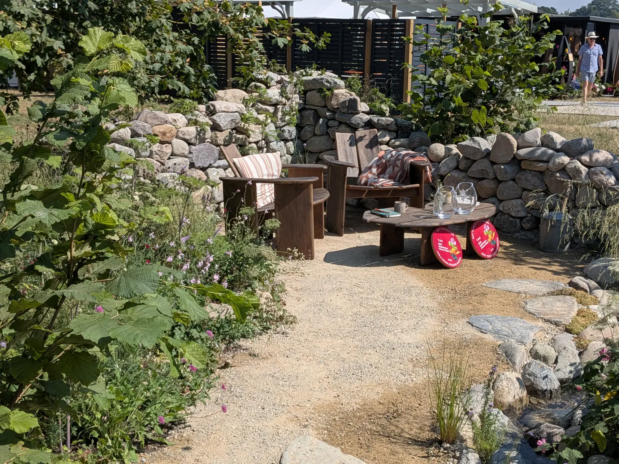 A tranquil Young Designer Garden showcases dry-stone walls, rustic wooden chairs and table on a gravel path, and a rock-lined rill flowing through naturalistic planting.