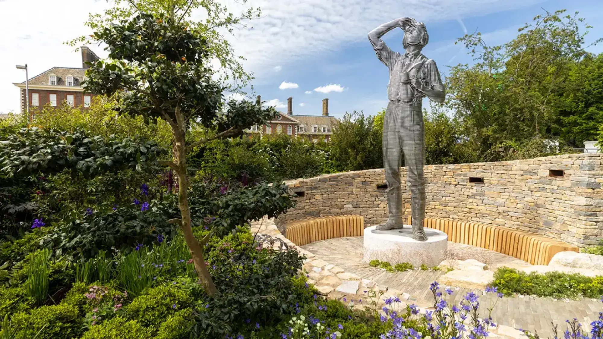 A wireframe sculpture of a pilot stands on a stone plinth in a show garden with a curved dry-stone wall, a timber bench, and naturalistic purple and green planting.