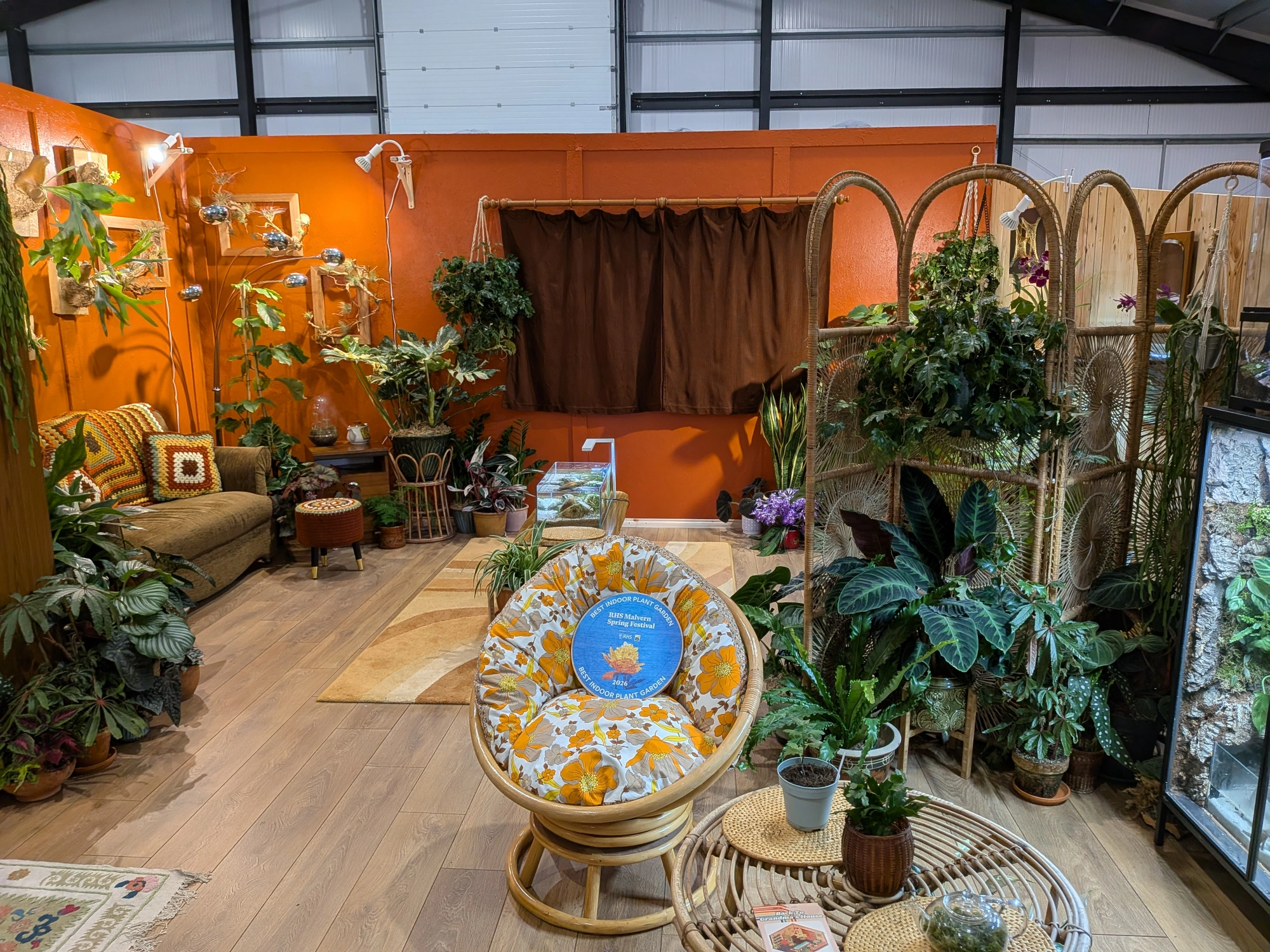 A cozy indoor plant garden featuring retro rattan furniture, a brown sofa with patterned cushions, and numerous lush green houseplants on a warm orange wall backdrop.