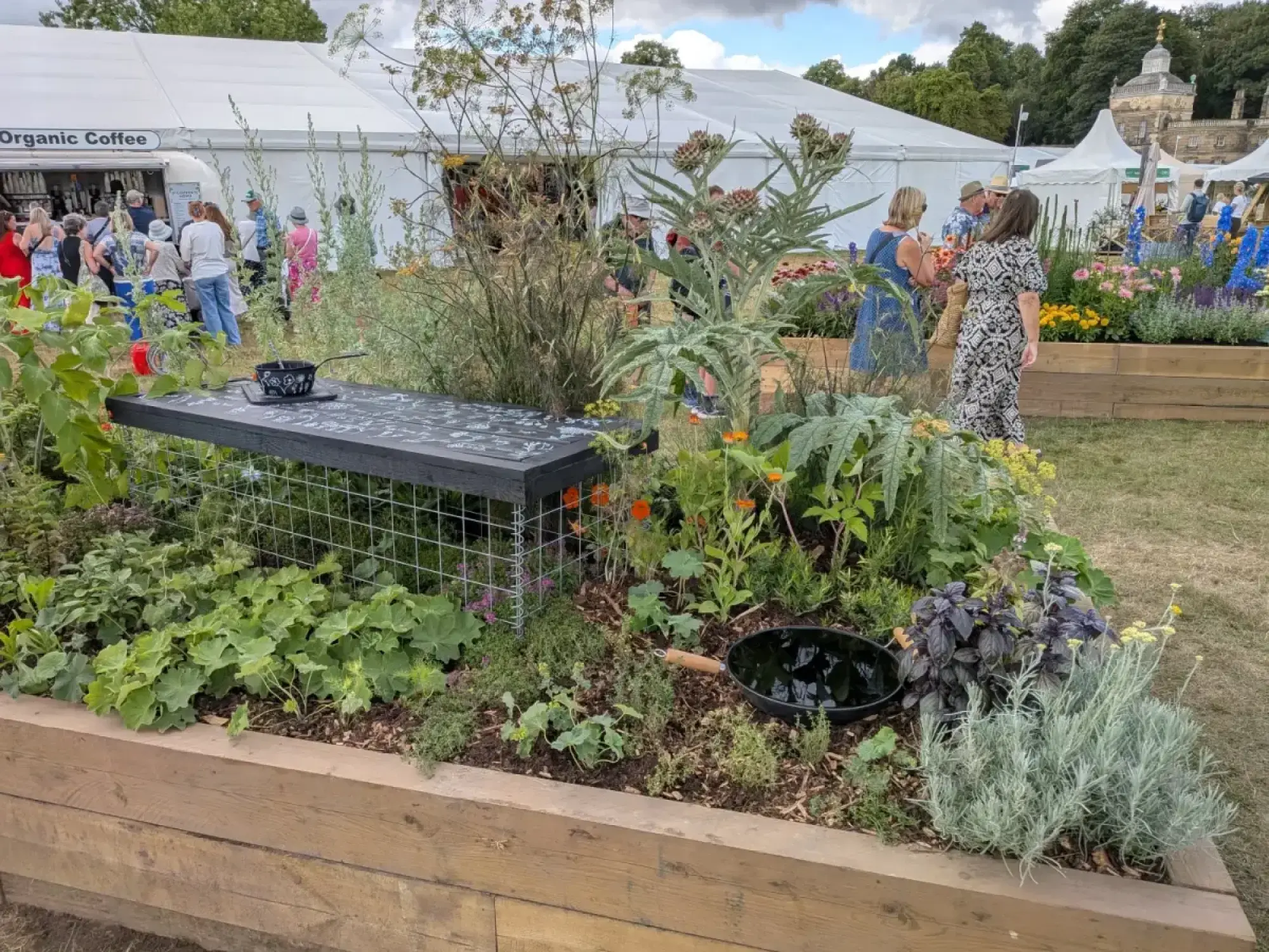 A long raised wooden border presents an 'Edimental Kitchen Classroom' with diverse edible plants, a black chalkboard bench, and a frying pan.