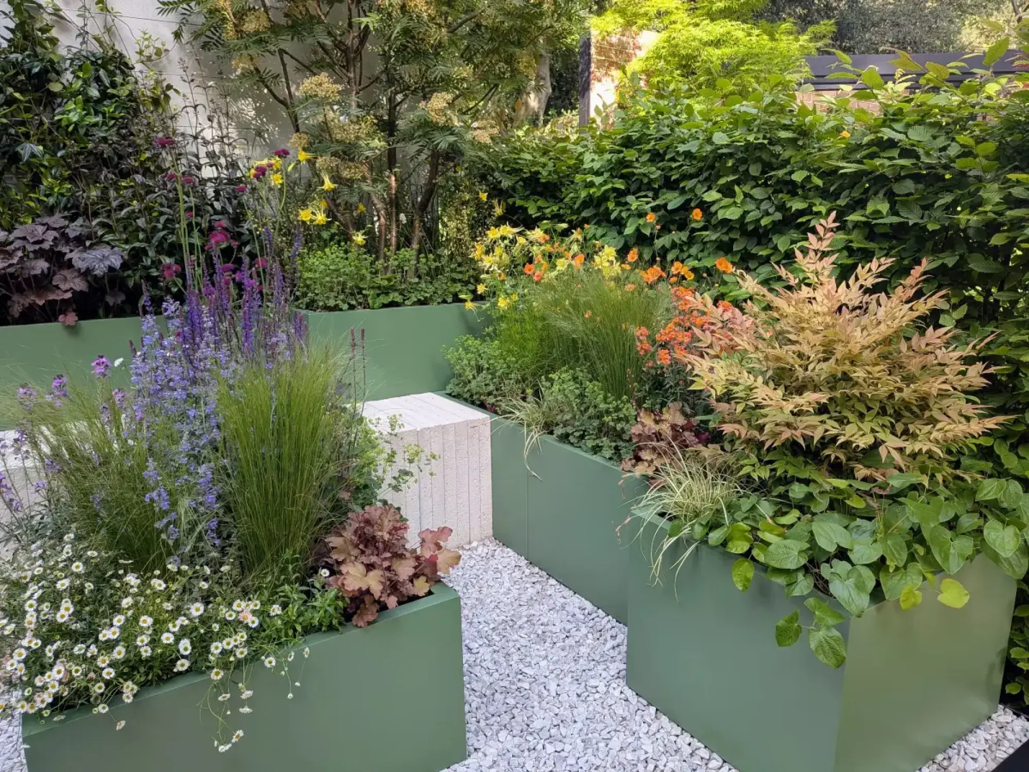 A contemporary container garden with sage green metal planters, a white stone bench, a gravel path, and diverse colourful flowers and grasses.