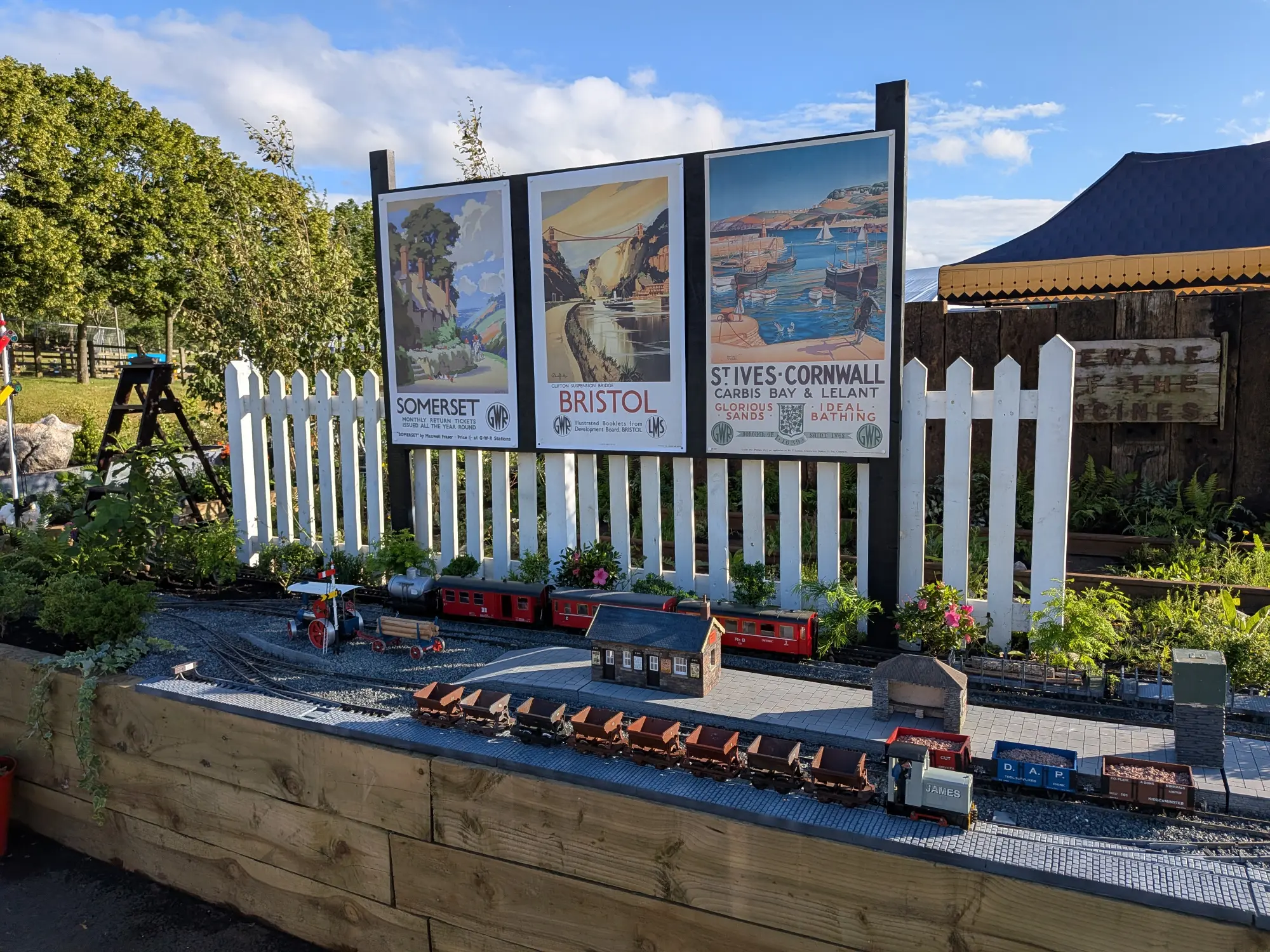 A charming garden features a detailed model railway with miniature trains, station models, a white picket fence, and nostalgic railway posters.