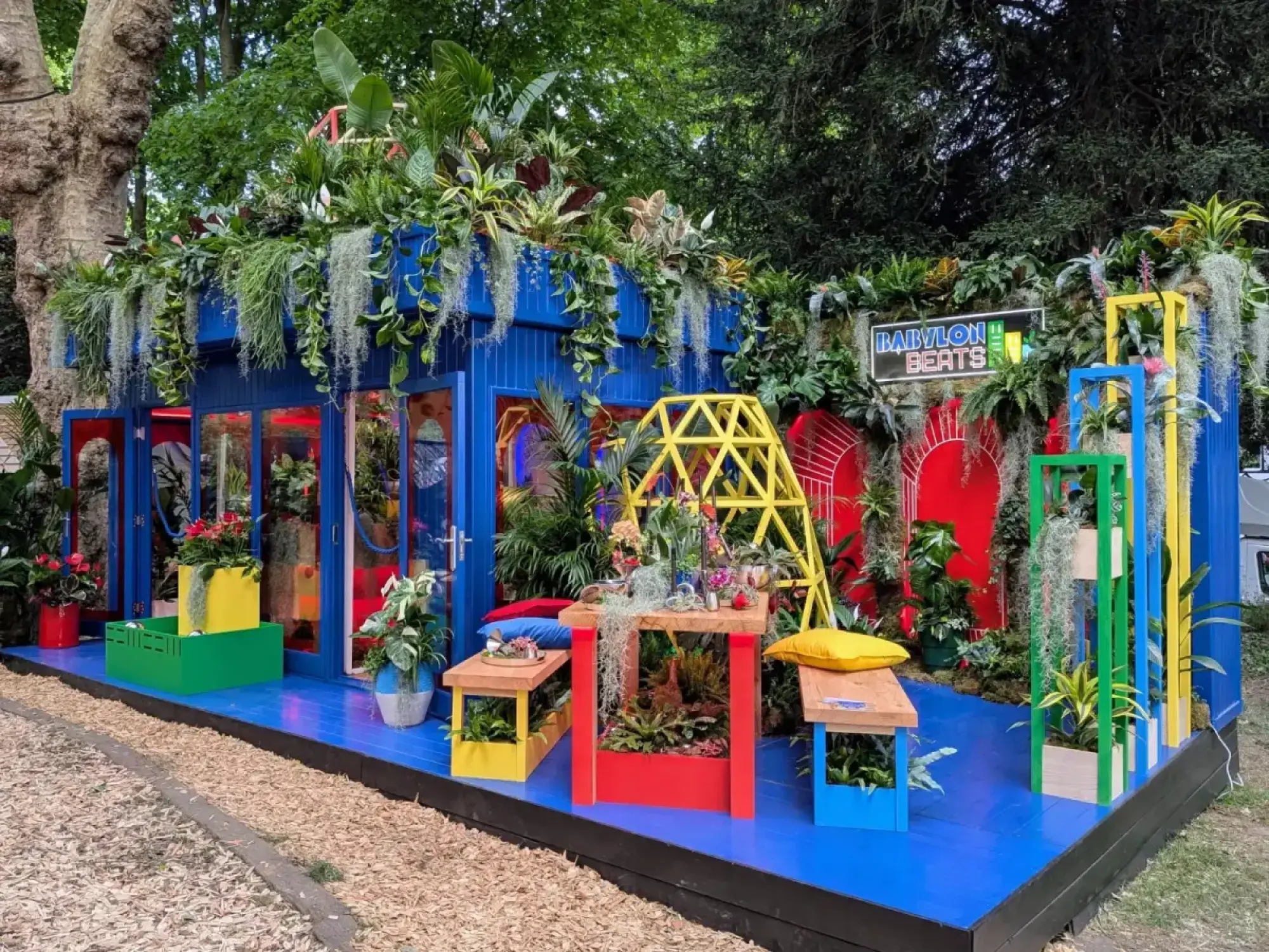 A vibrant houseplant studio with a royal blue facade, featuring abundant tropical plants, colourful red, yellow, and green furniture, and a bright neon sign.