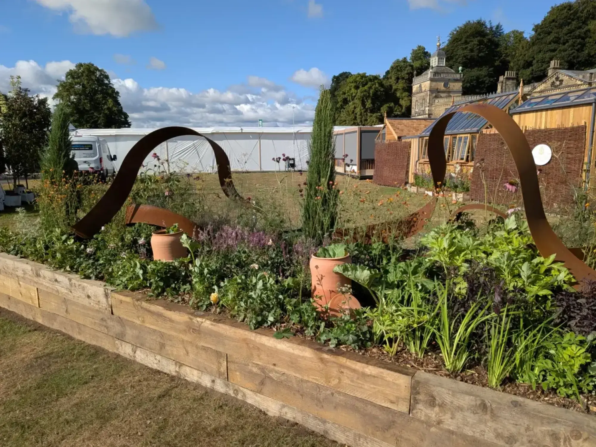 A raised timber border displays curving rust-coloured metal ribbons, dense botanical planting with purple flowers and green foliage, and terracotta pots.