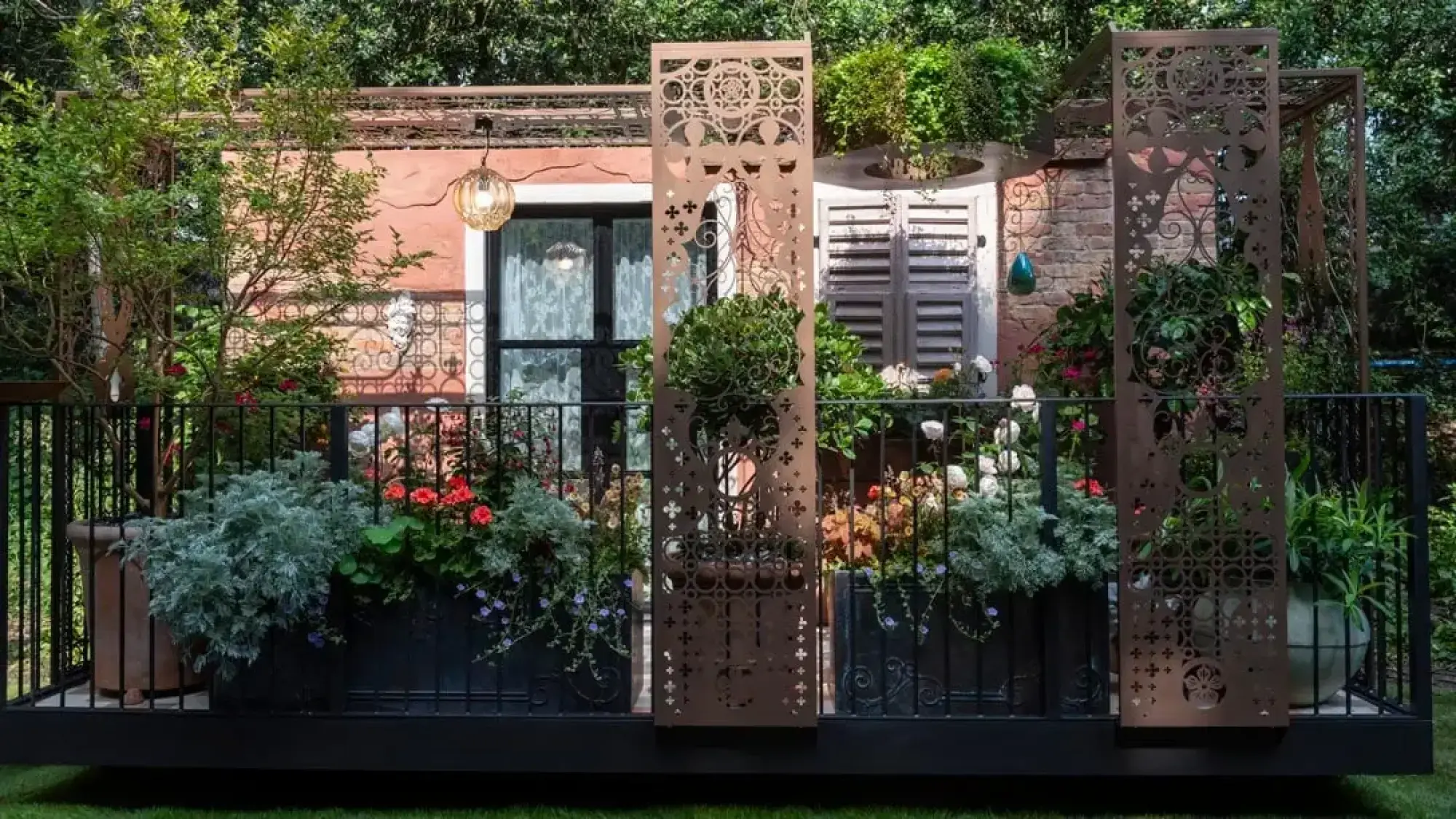A romantic balcony garden featuring ornate laser-cut metal screens, lush potted plants including red geraniums and white roses, and a detailed black wrought iron railing against a terracotta-colored facade with shutters.