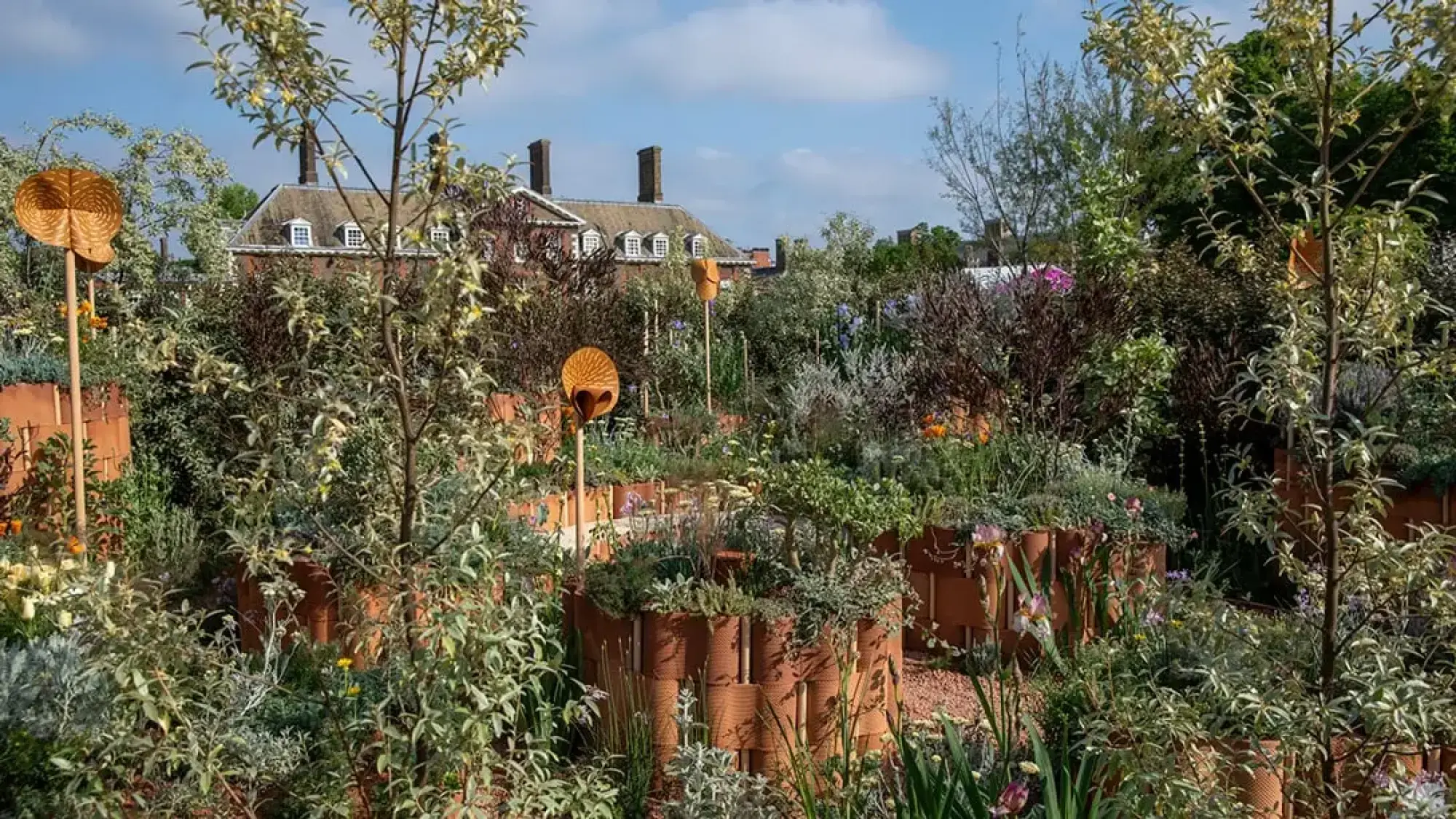 A sanctuary garden with circular woven terracotta planters and tall sound mirrors, displaying naturalistic planting, small trees, and orange, purple, yellow flowers.
