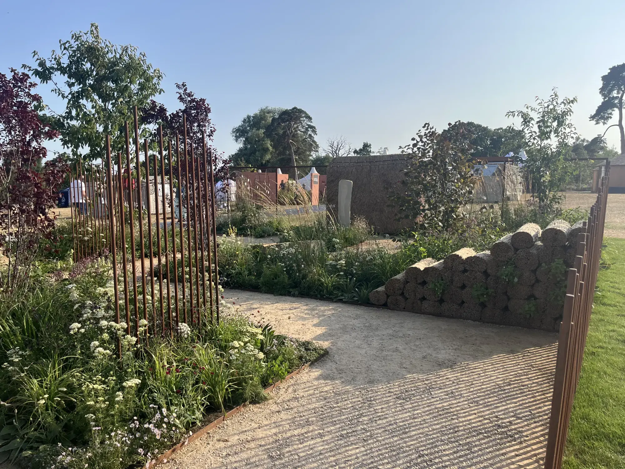 A naturalistic garden with a gravel path, a rustic wall of stacked organic bales, corten steel rod screens, and beds of wildflower planting.
