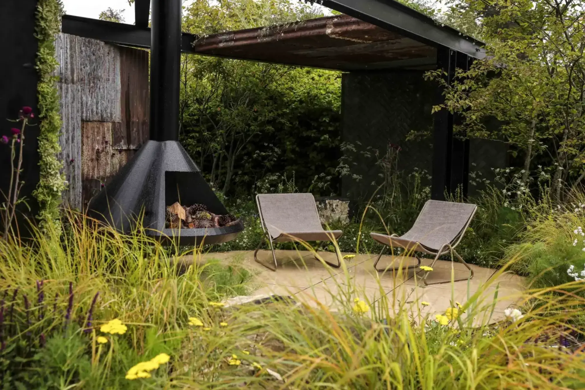 A contemporary garden features a black conical fireplace, two beige lounge chairs on a light patio, under a rusted metal canopy, with wild grasses.