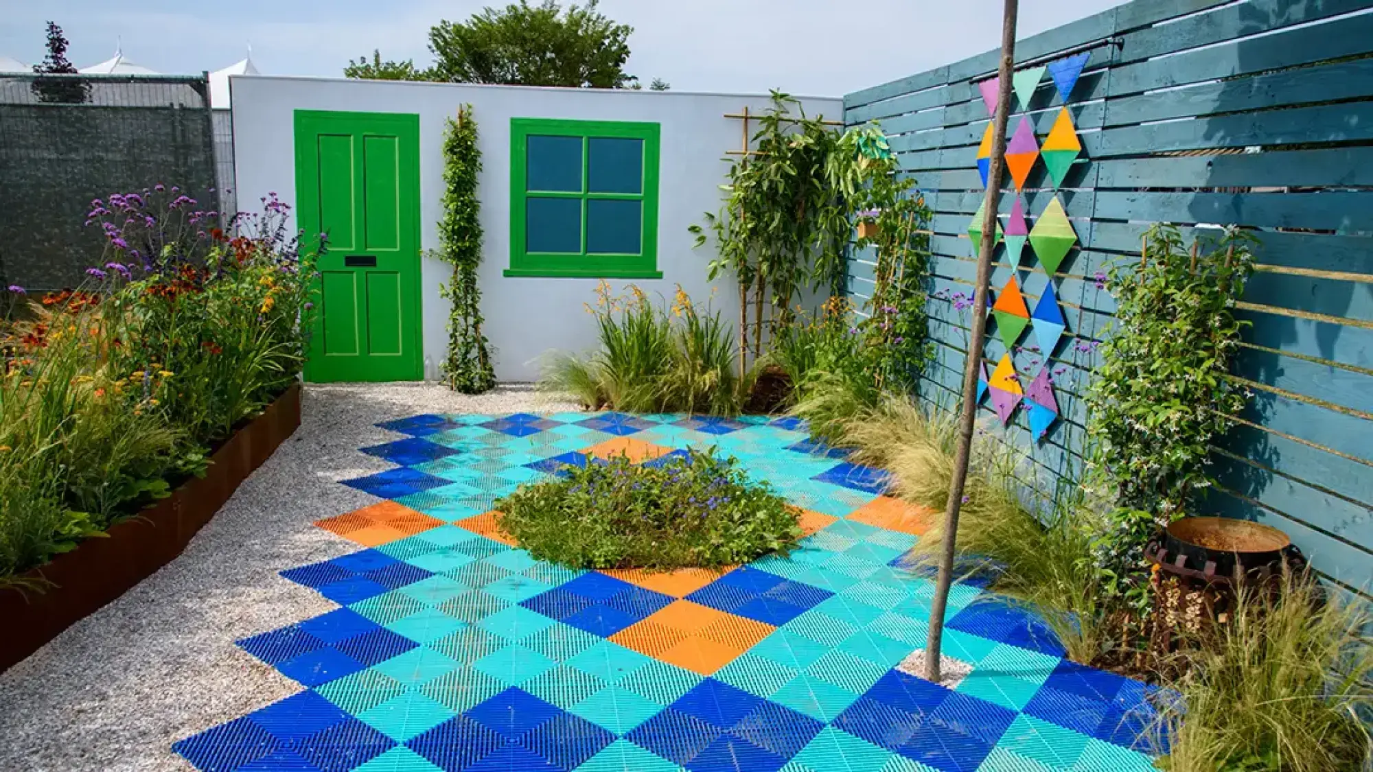 A vibrant front garden with a geometric patterned patio of blue, aqua, and orange tiles leading to a bright green door and window on a white wall, bordered by a blue slatted fence with diamond art and lush plantings.