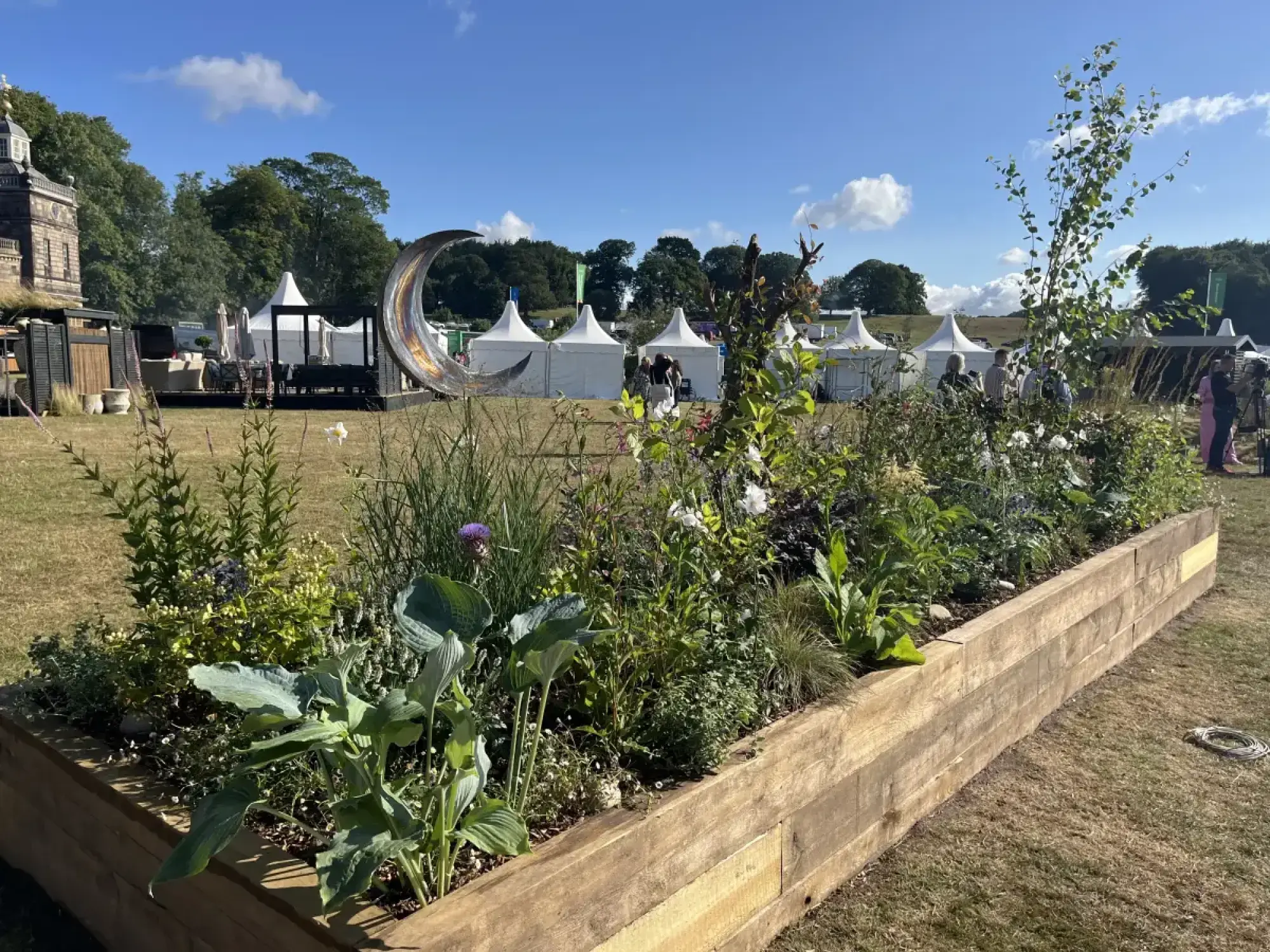 A long raised garden border with mixed planting of white and purple flowers, and a metallic crescent moon sculpture.