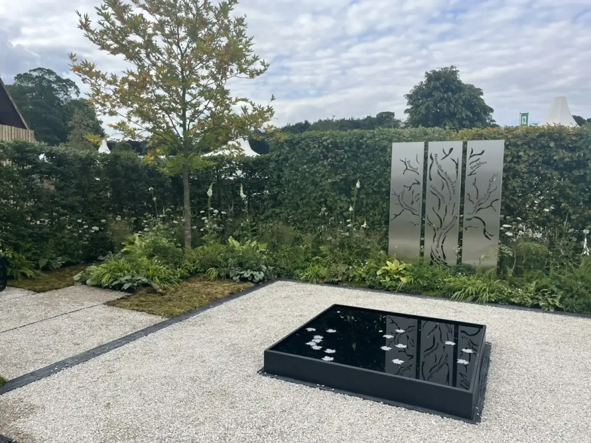 A contemporary show garden features three tall metal tree panels, a dark reflective water feature with white floating leaves, and lush green planting with white flowers.