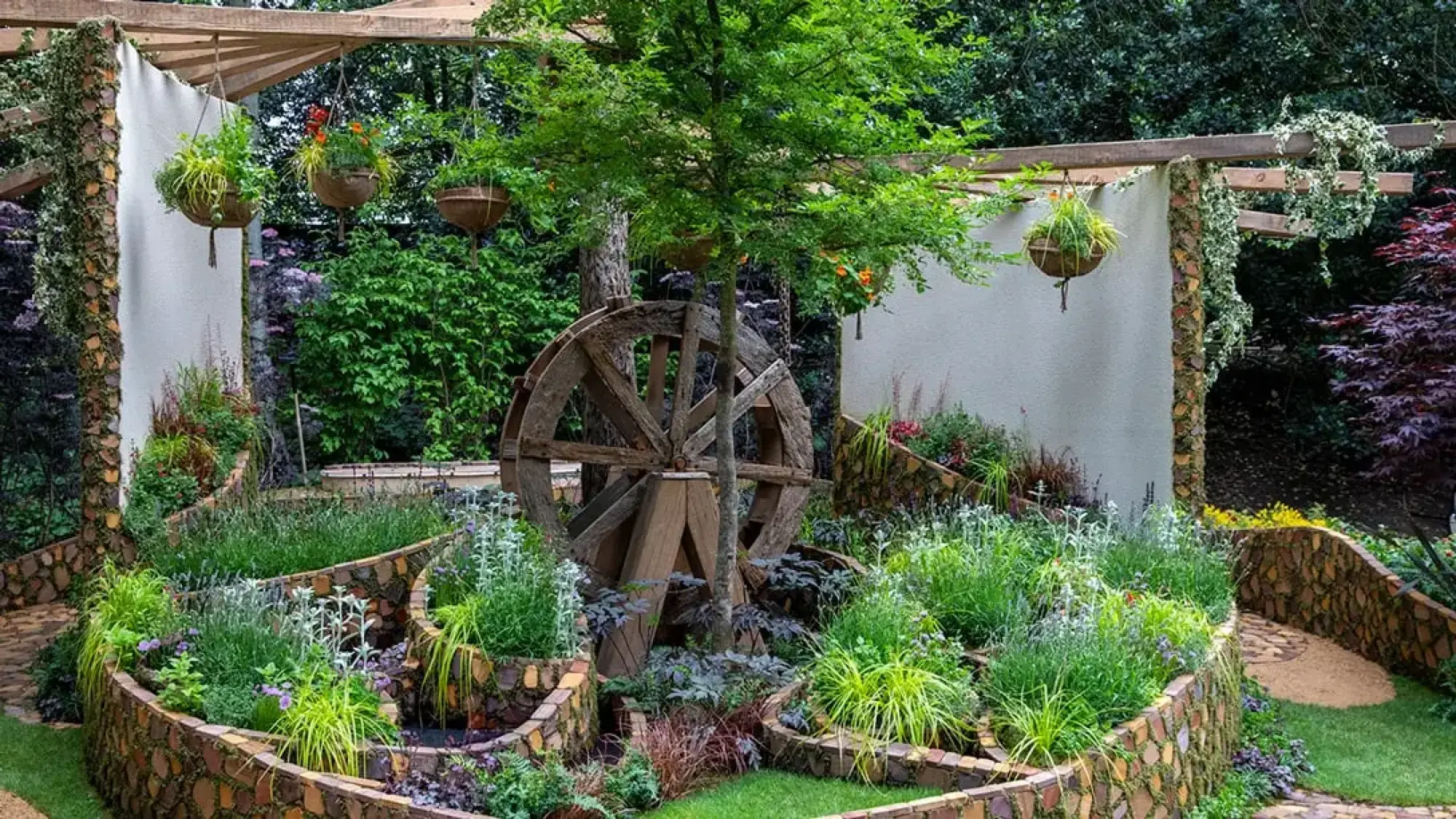 A naturalistic sanctuary garden features a large wooden water wheel and tiered, wood-faced planters overflowing with green and purple plants and a winding path.