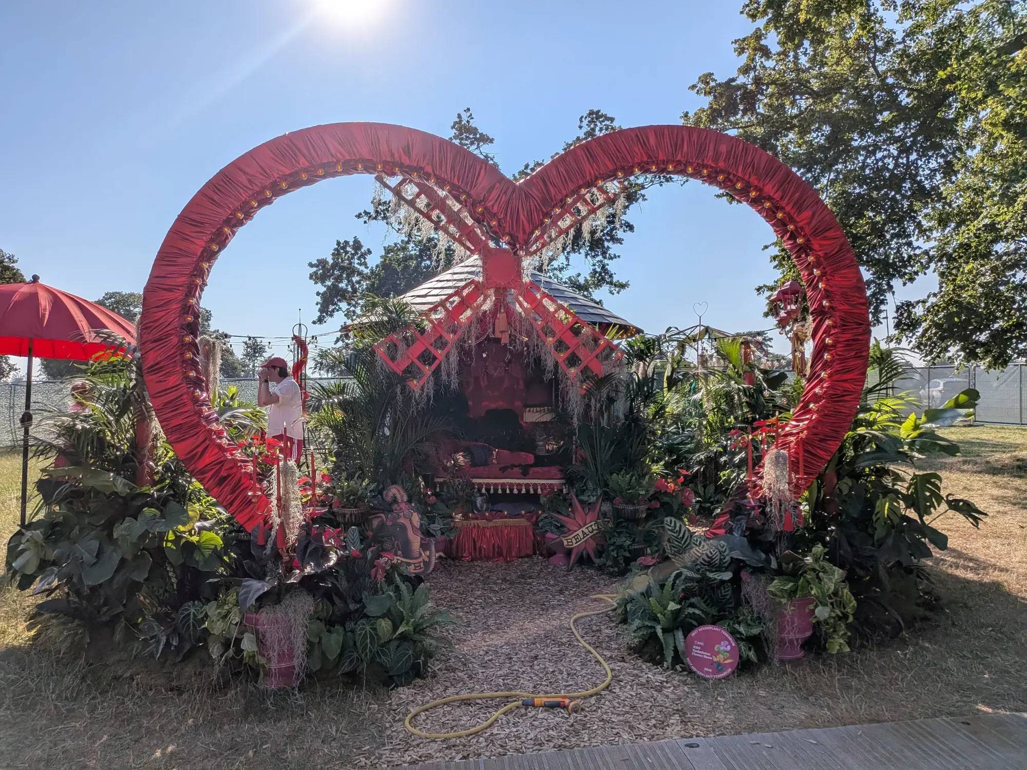 A lush, opulent houseplant display features a giant red heart-shaped archway framing a central draped seating area with tropical plants.
