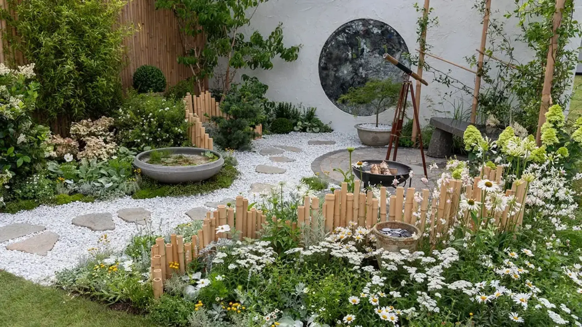 A tranquil Japanese-style garden featuring a white gravel path, lush green and white planting, a round window in a white wall, a water bowl, and a telescope near a fire pit.