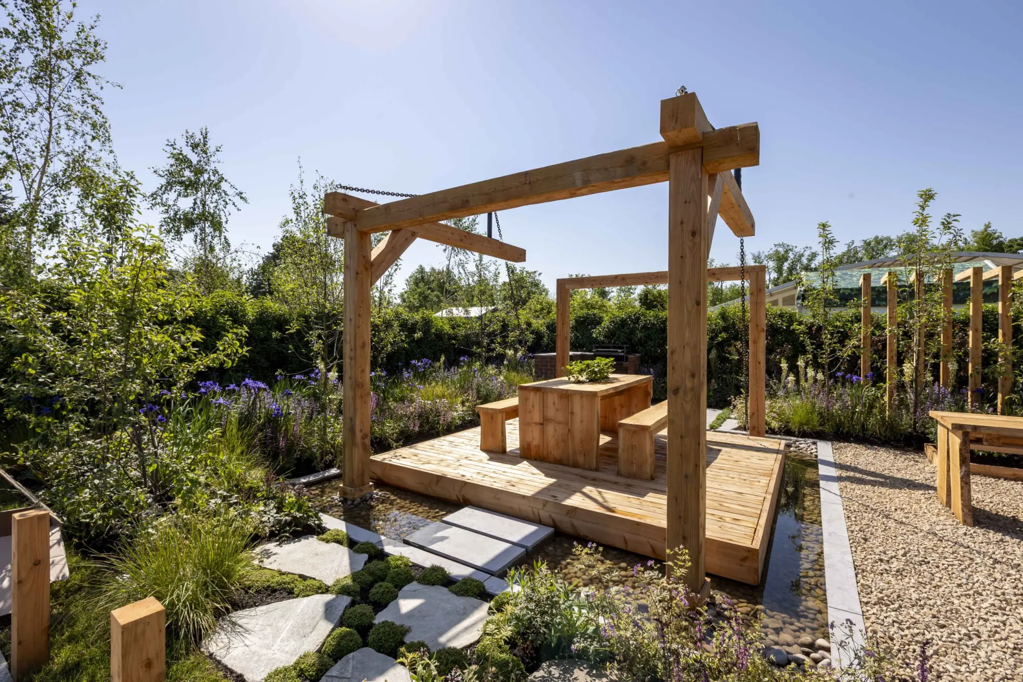 An elevated timber deck with a picnic table and benches is surrounded by a water rill and naturalistic planting, featuring purple irises and a timber pergola.
