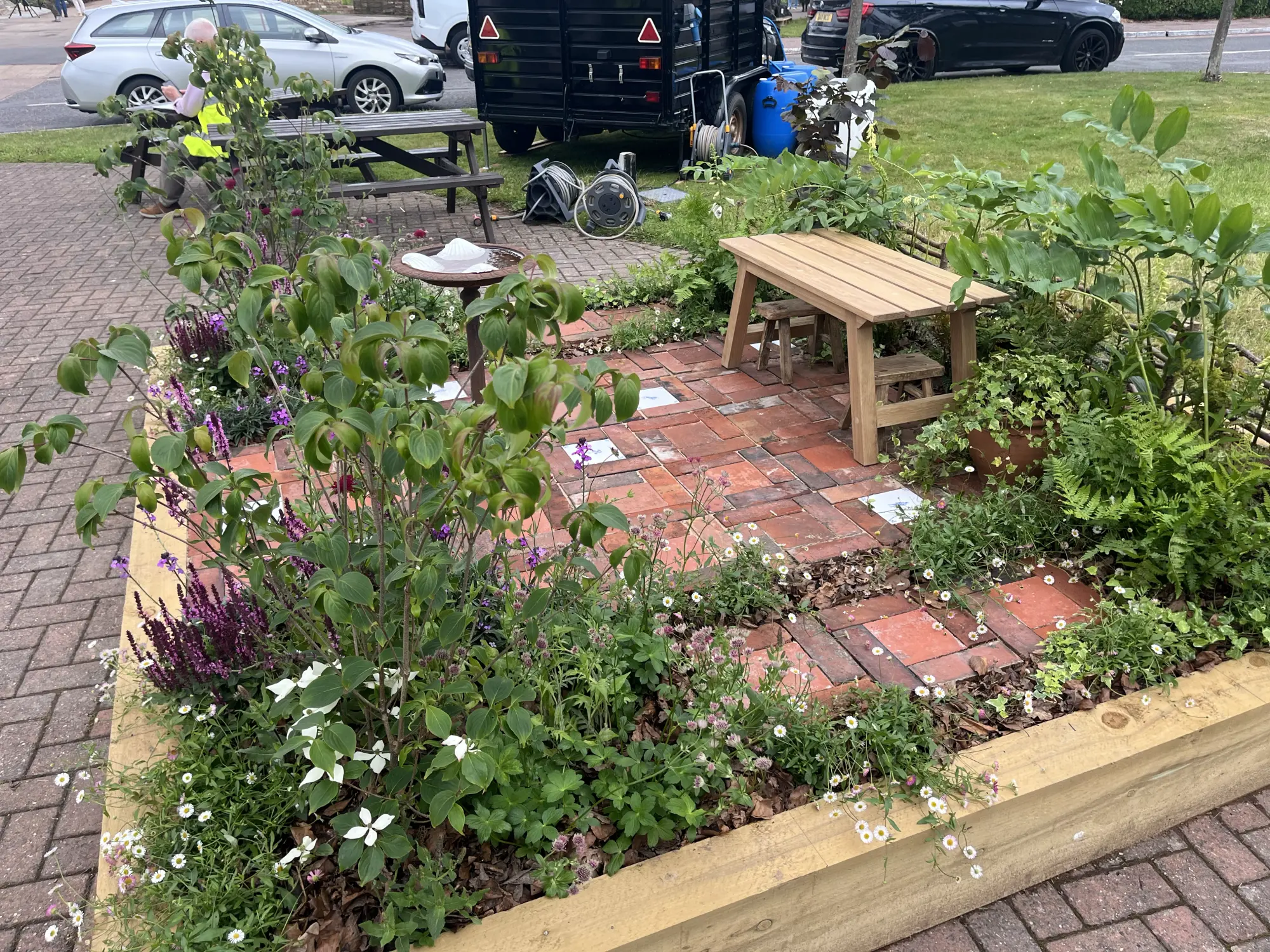 A beautiful border garden features a light wood picnic table on red brick paving, surrounded by a timber raised bed abundant with green plants and white, purple, and pink flowers.