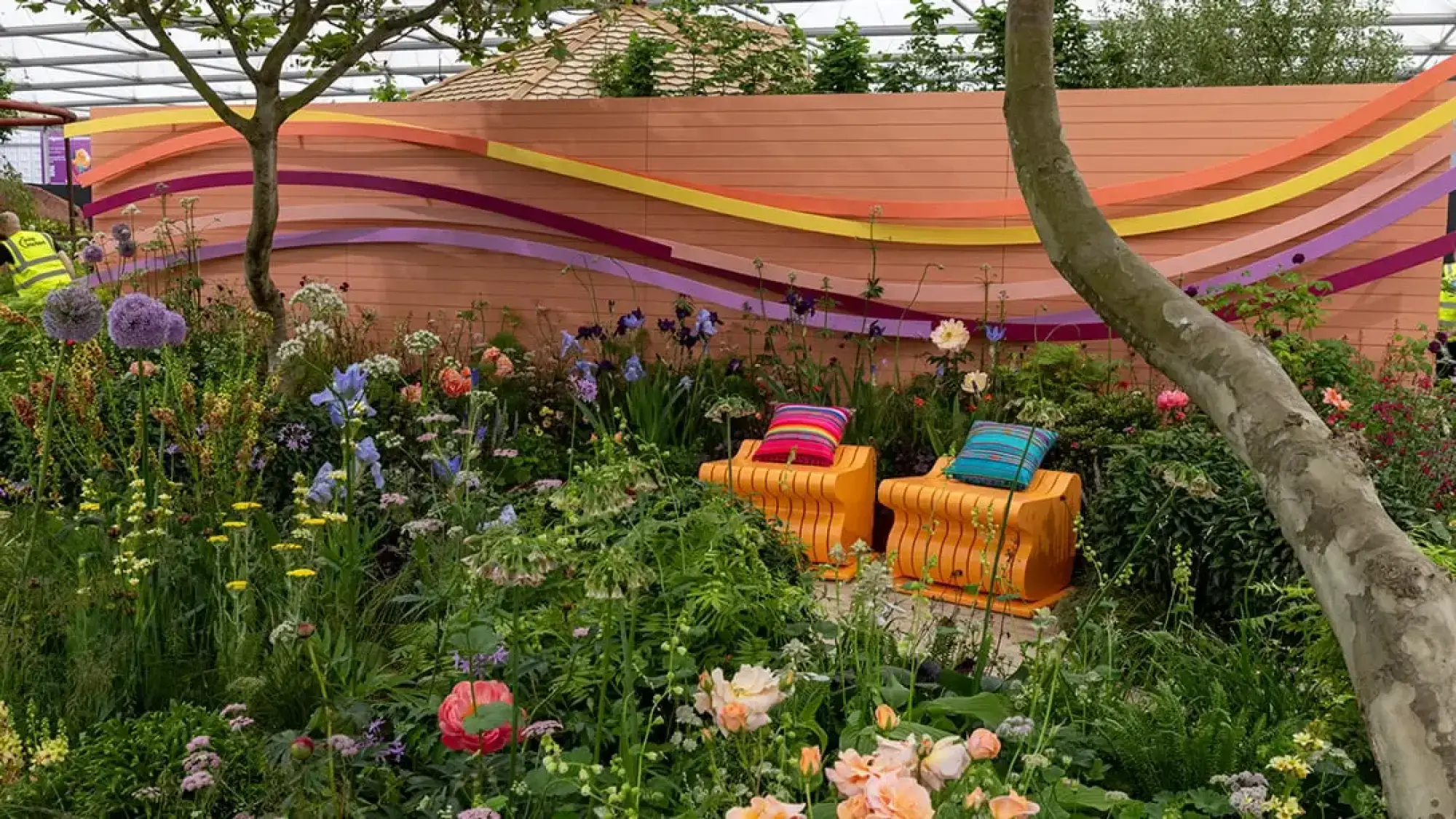 The Panathlon Joy Garden has orange slatted benches, a peach wall with wavy colourful ribbons, and abundant, vibrant mixed flowers.