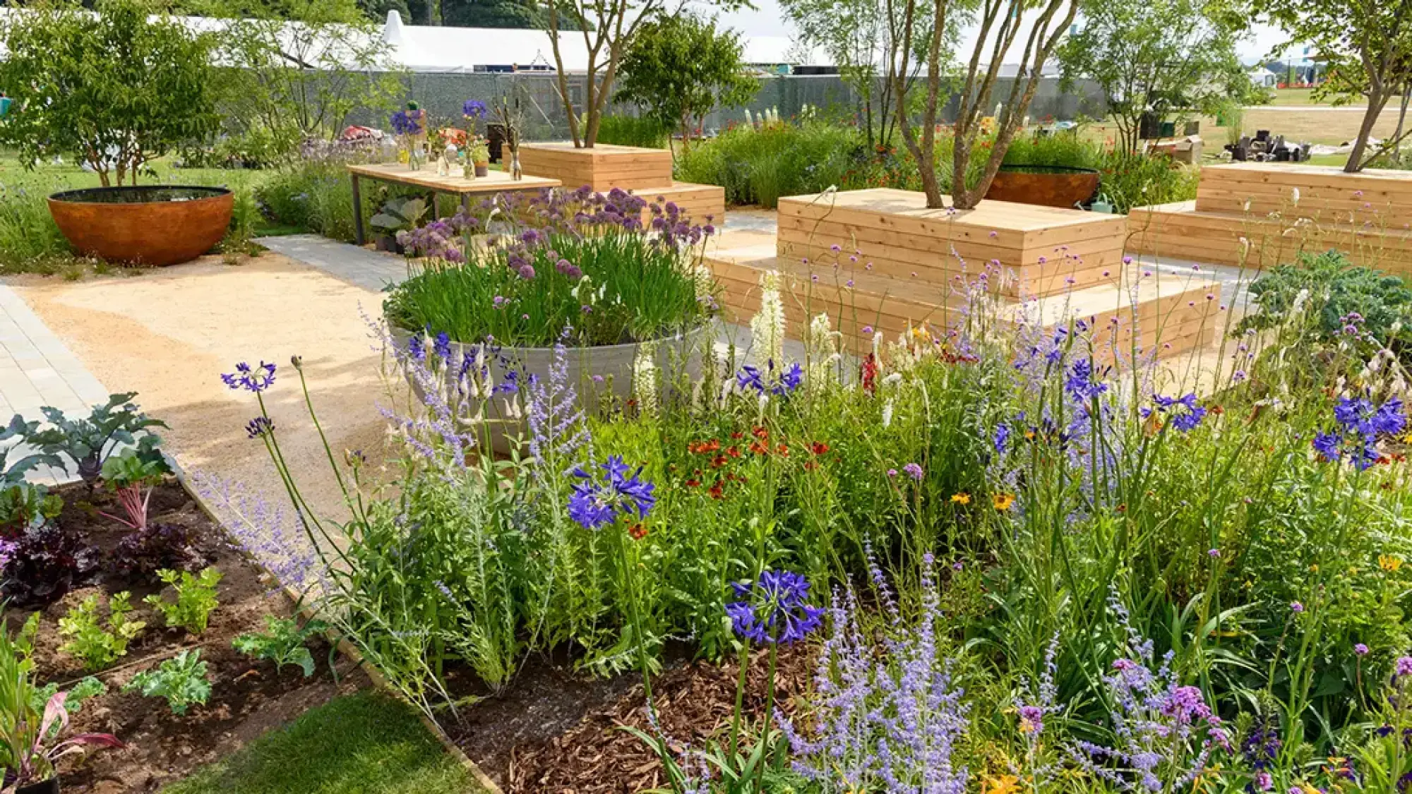 A contemporary feature garden with tiered light wood planters, a rusted water bowl, a wooden dining table, and raised beds filled with colourful perennial flowers and edible plants alongside gravel paths.