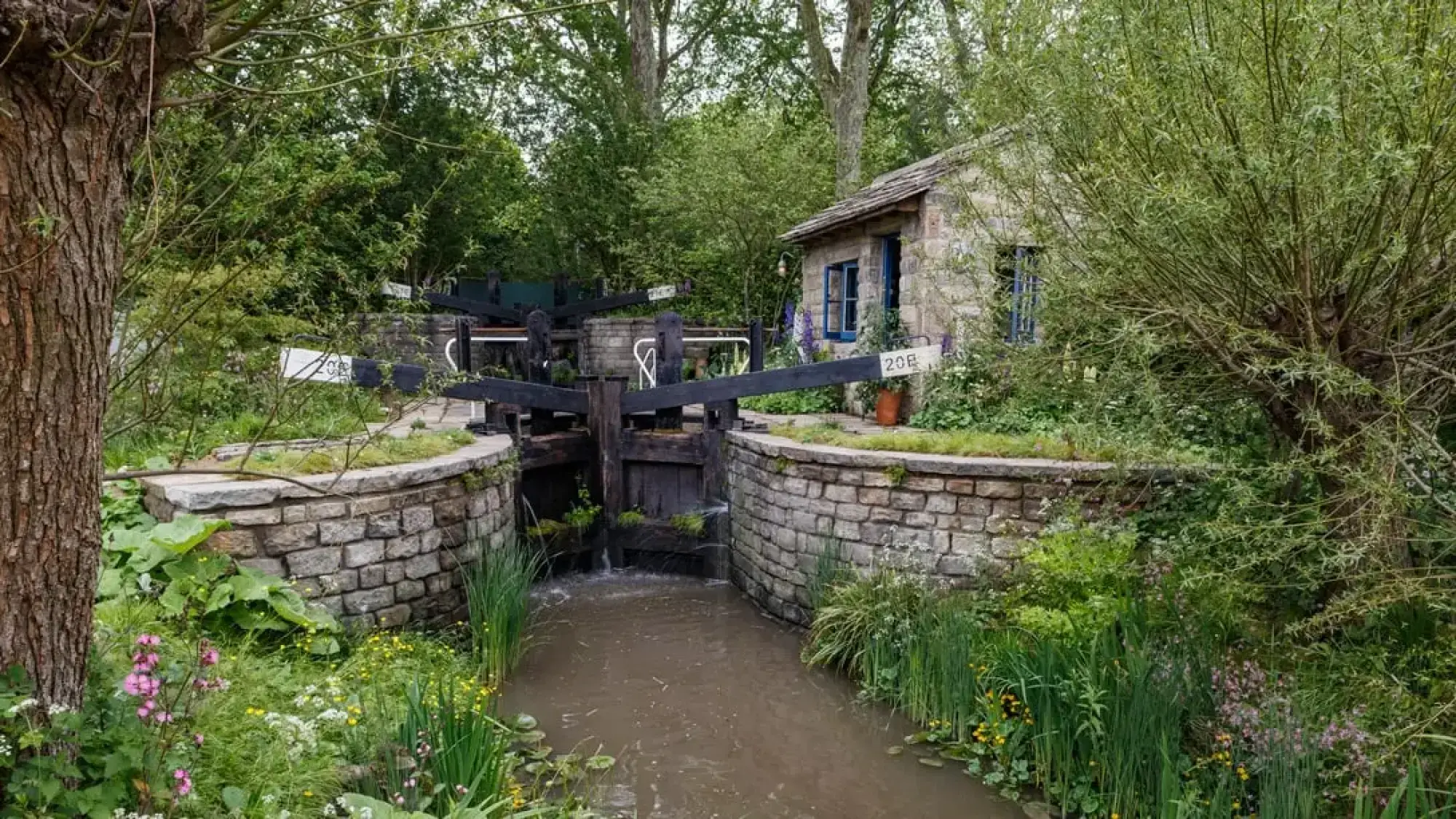 A rustic garden features a stone cottage with blue windows, black wooden canal lock gates, a flowing water channel, and lush planting.